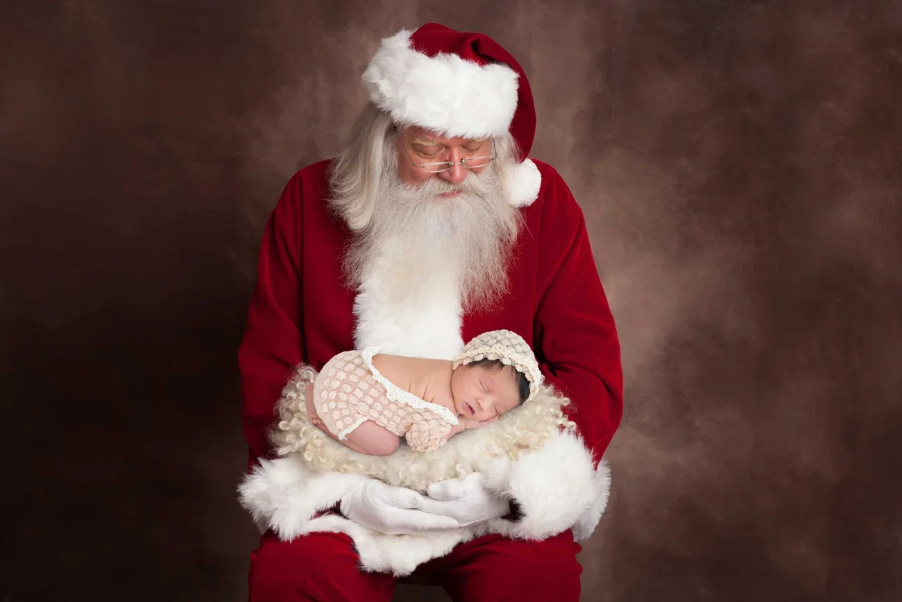 Santa Claus with glasses and a red suit holding a newborn sleeping on a fluffy white blanket against a brown background.