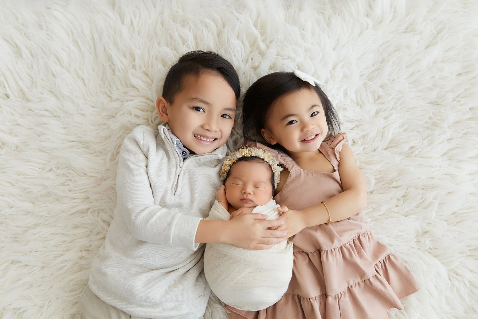 A young boy and girl with their newborn sibling lying on a cream-colored fluffy rug.