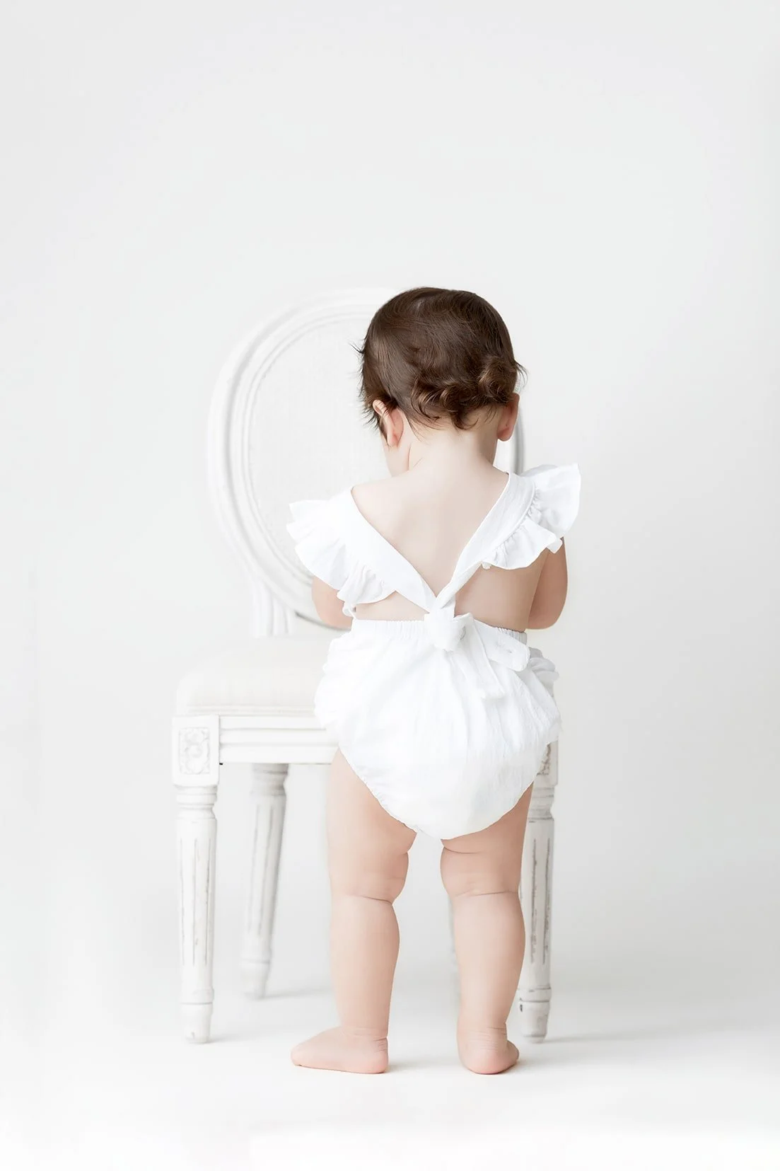 Child with short brown hair wearing a white outfit, seen from the back, standing in front of a white vanity with a round mirror.
