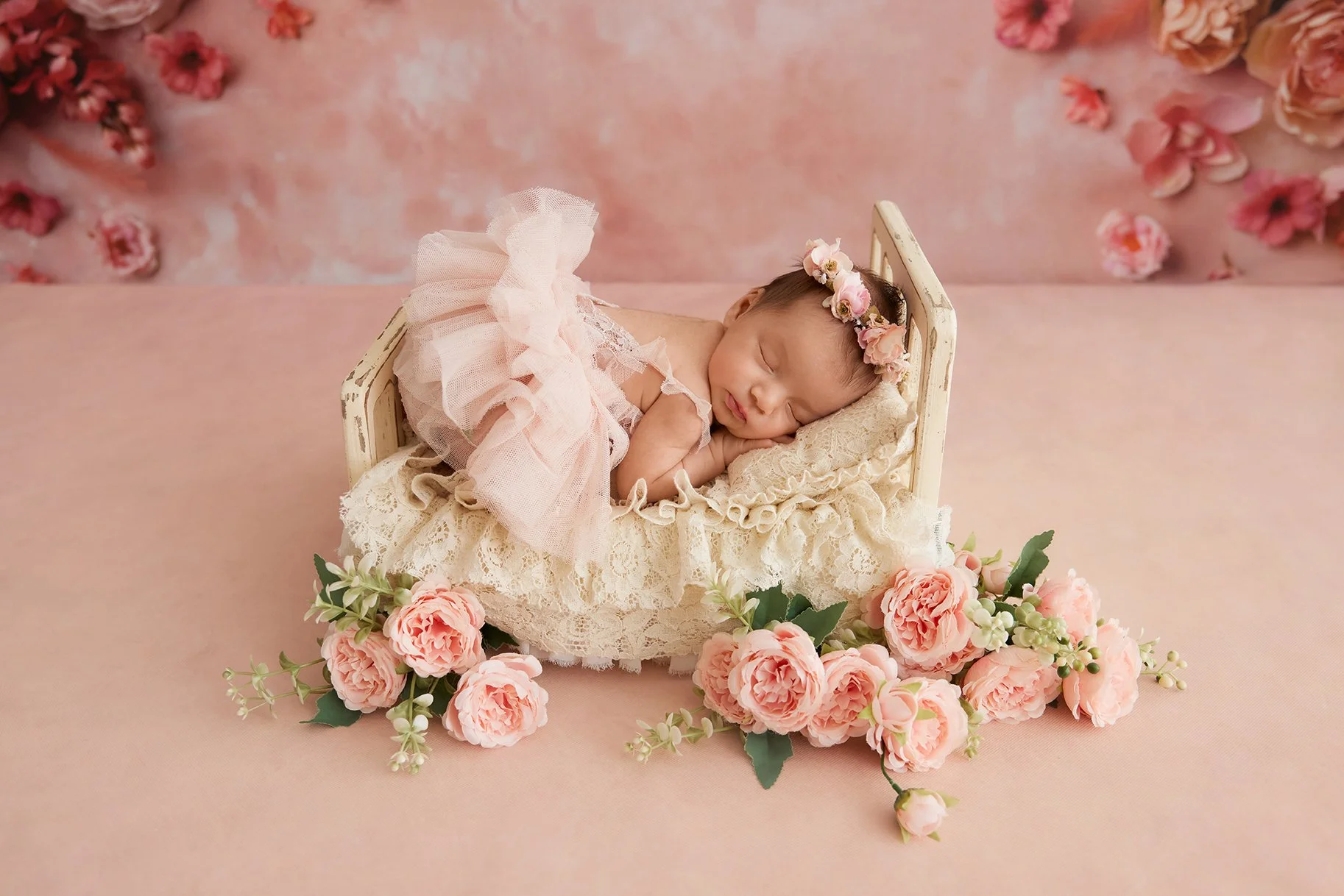 A sleeping newborn baby girl wearing a pink frilly dress and a floral headband, lying on a lace pillow in a small wooden cradle surrounded by pink roses and green leaves, with a pink floral backdrop