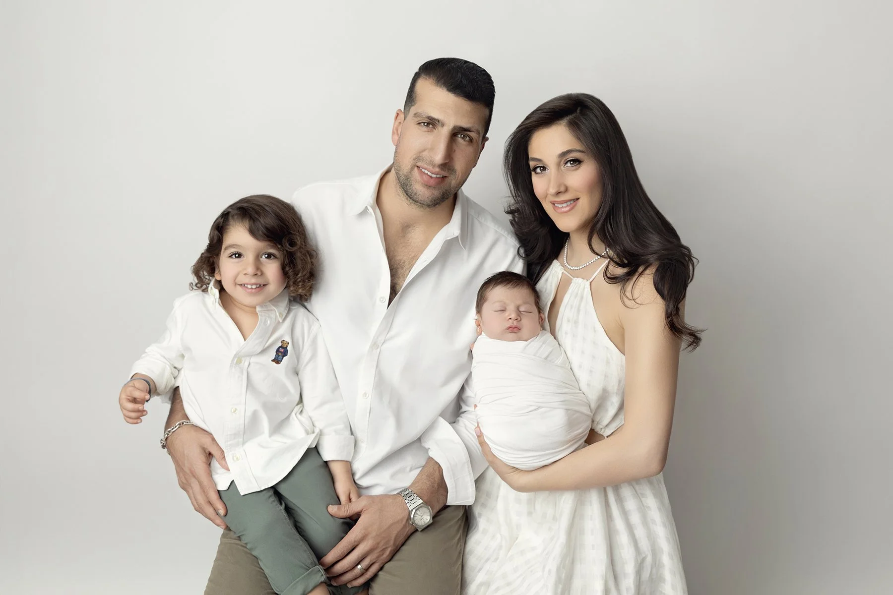 Family portrait of four members: a father, mother, and two children, one boy and one newborn, standing against a plain light gray background.