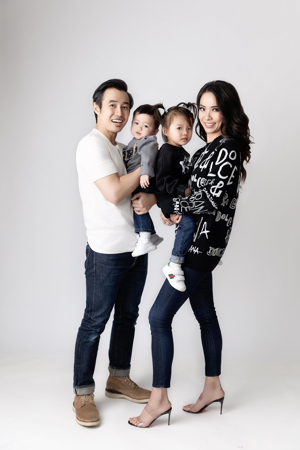 A happy family of four posing together against a plain white background. The father, dressed in a white t-shirt and jeans, is holding a young boy. The mother, in a black graphic sweater and high heels, is holding a young girl. Both children are dress