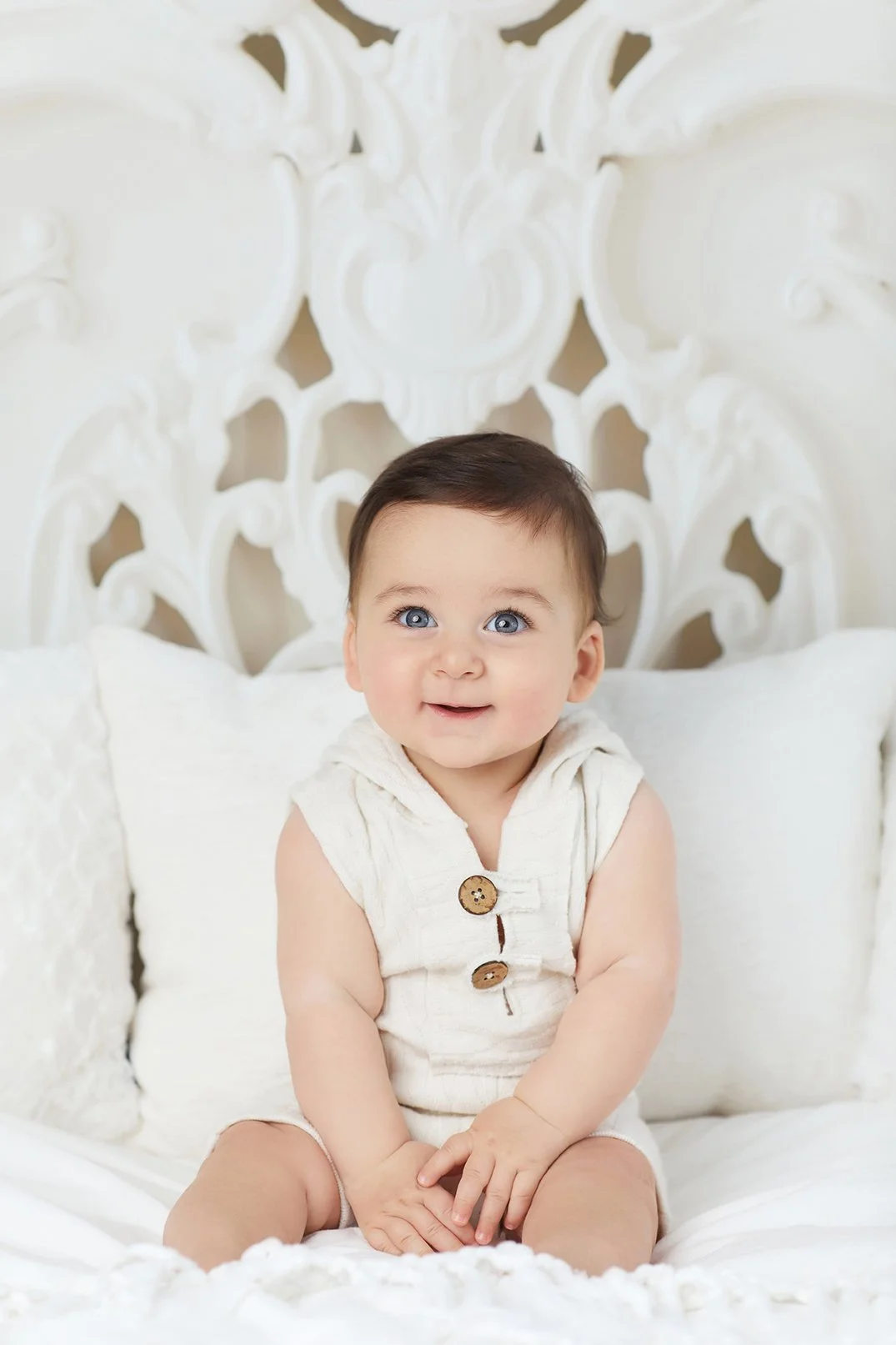A smiling baby with blue eyes and dark hair, sitting on a white bed with white pillows and an intricate white headboard in the background.