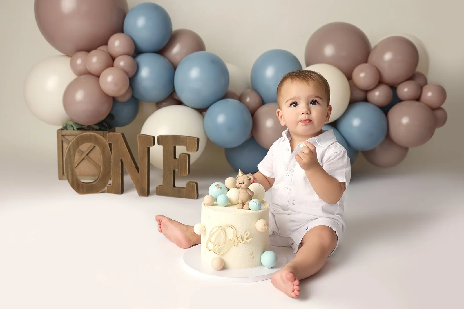 A baby celebrating their first birthday with a cake, balloons, and decorative letters behind them spelling 'ONE'.