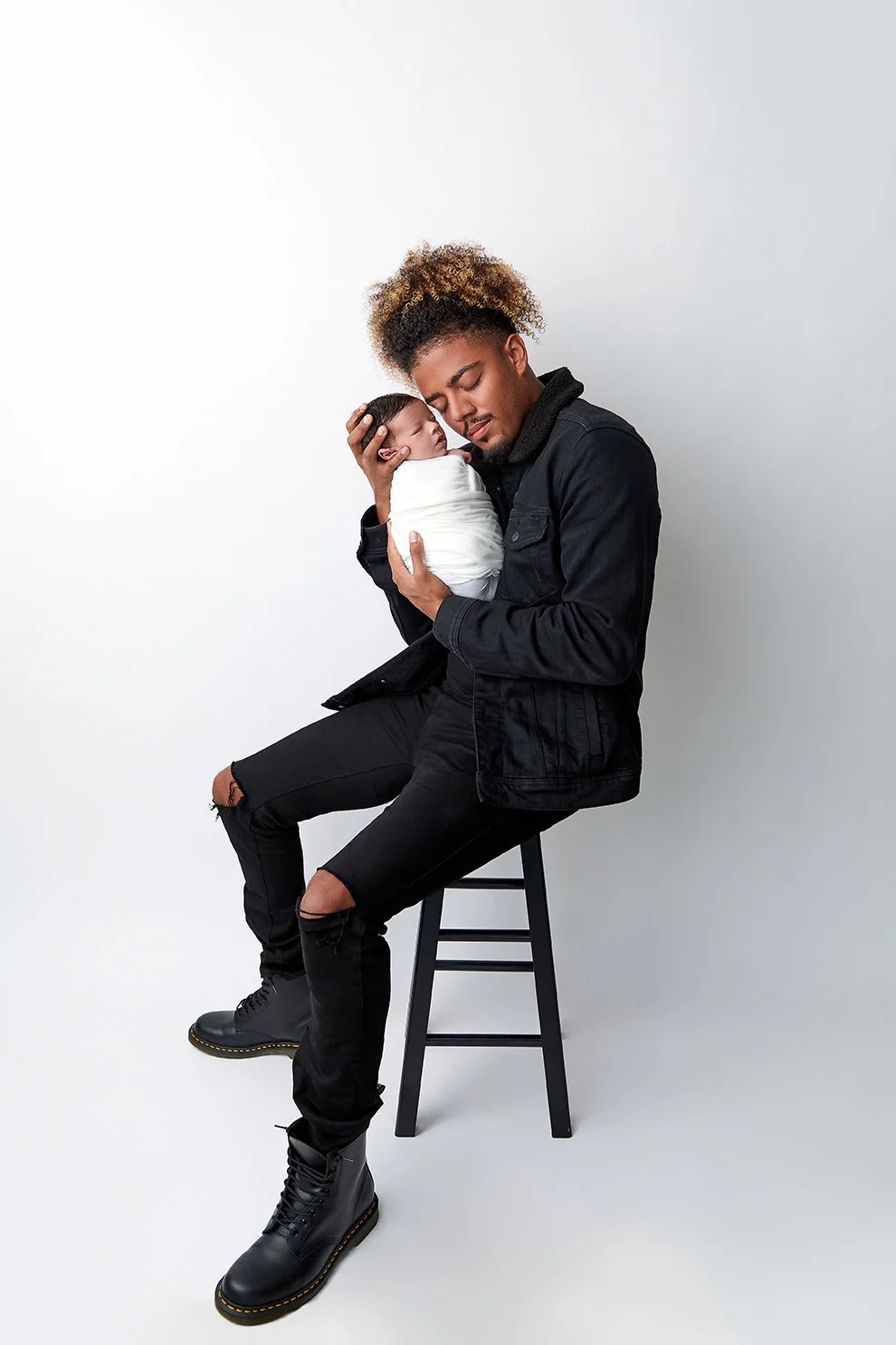 A man with curly hair and a beard holding a sleeping newborn wrapped in a white blanket, sitting on a stool against a plain light gray background.
