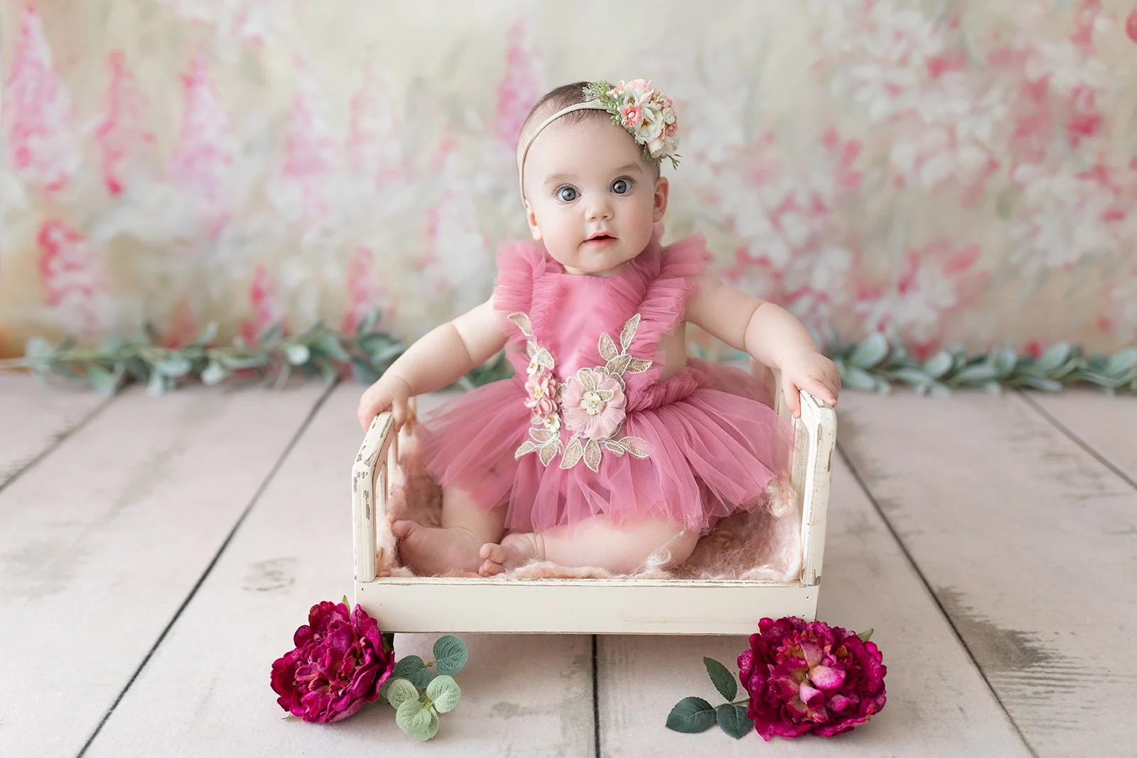 A baby girl in a pink tulle dress with floral embroidery sits in a small distressed white wooden bed, surrounded by pink peonies and green foliage, with a floral backdrop.