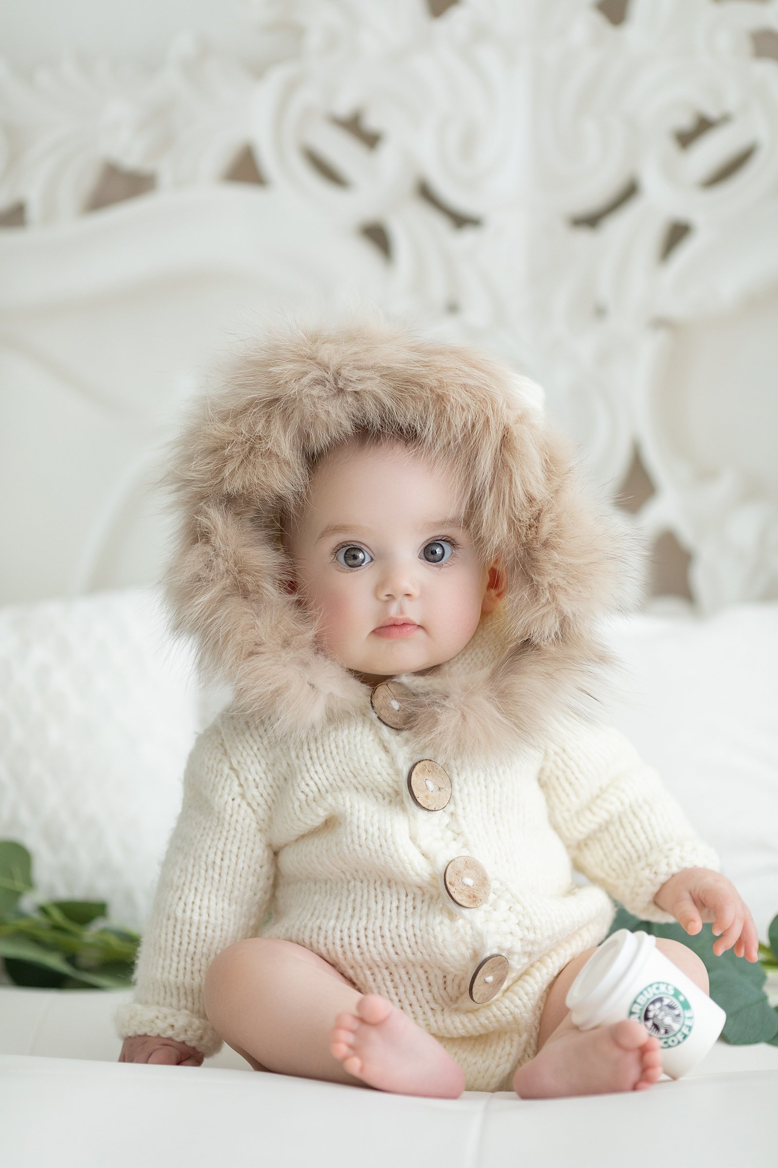 A baby sitting on a white surface wearing a cream-colored knitted sweater with buttons and a large furry hood, holding a Starbucks cup, with a white ornate headboard in the background.