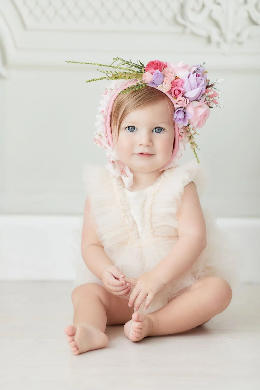 A baby girl with blue eyes, sitting on a white floor, wearing a white dress and a floral bonnet decorated with pink, purple, and white flowers and green foliage.