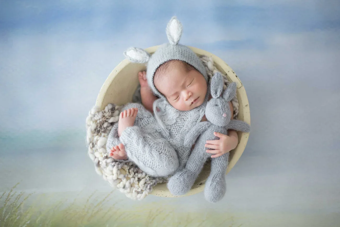 A sleeping baby dressed in gray knitted clothing, holding a small gray stuffed bunny, and wearing a gray hat with bunny ears, lying in a round basket on a fluffy blanket.