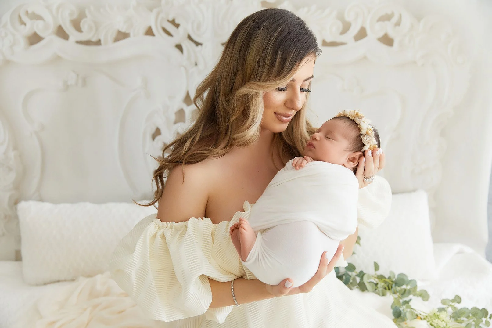 A woman with long wavy brown hair holding a sleeping baby wrapped in white cloth, wearing a floral headband, in a bright room with ornate white decor.