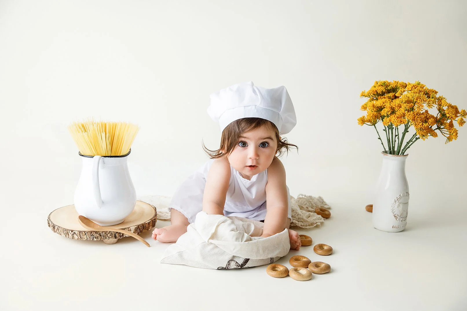 Baby dressed as a chef in a white apron and chef hat, surrounded by food items including dried bagels, spaghetti in a jug, and a vase of yellow flowers, on a white background.