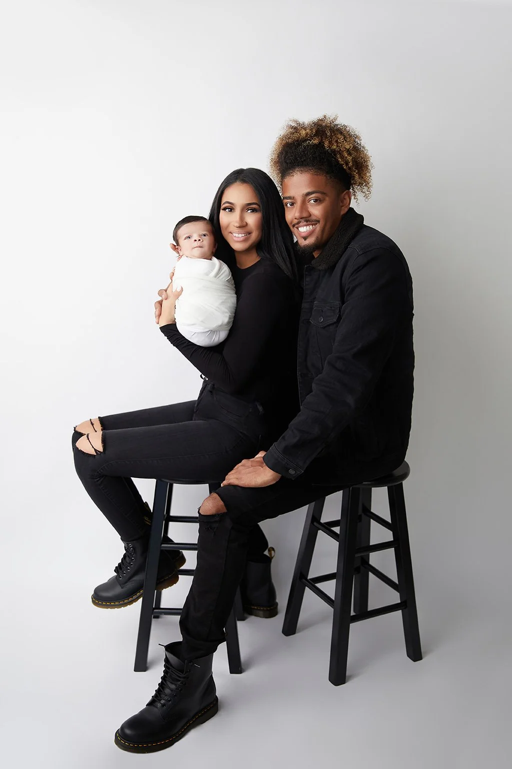 A young family sitting on two black stools against a plain white background. The woman holds a newborn wrapped in white, and the man is seated next to her. They are all smiling and dressed in black.