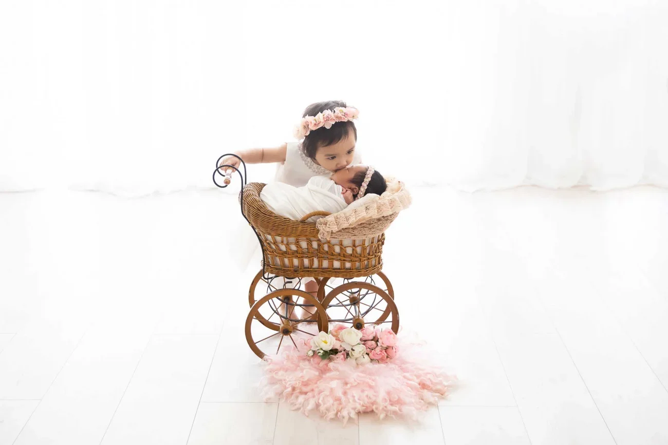 Two young children in a wicker toy stroller with pink flowers and soft pink rug, one girl with a flower crown is kissing the other girl who is lying down with a matching headband.