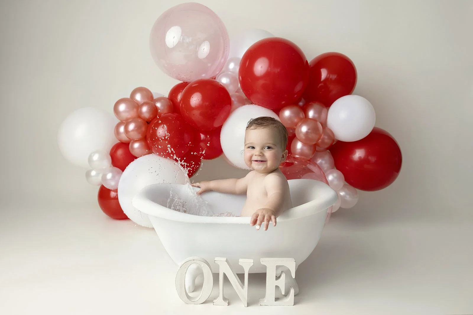 A smiling baby sitting in a white bathtub, surrounded by red, white, and pink balloons, with the word 'ONE' spelled out in white letters at the front, celebrating a first birthday.