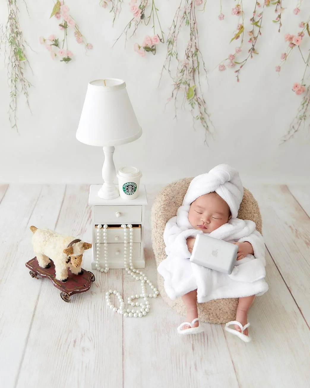 A sleeping baby wrapped in a white towel with a towel turban on her head, sitting in a cozy chair. The baby holds a small laptop. Next to her is a small white nightstand with a lamp, a Starbucks coffee cup, and a pearl necklace draped on the floor. A plush toy cow on a miniature wagon is nearby, and the background features a wall with pink flowers hanging down.