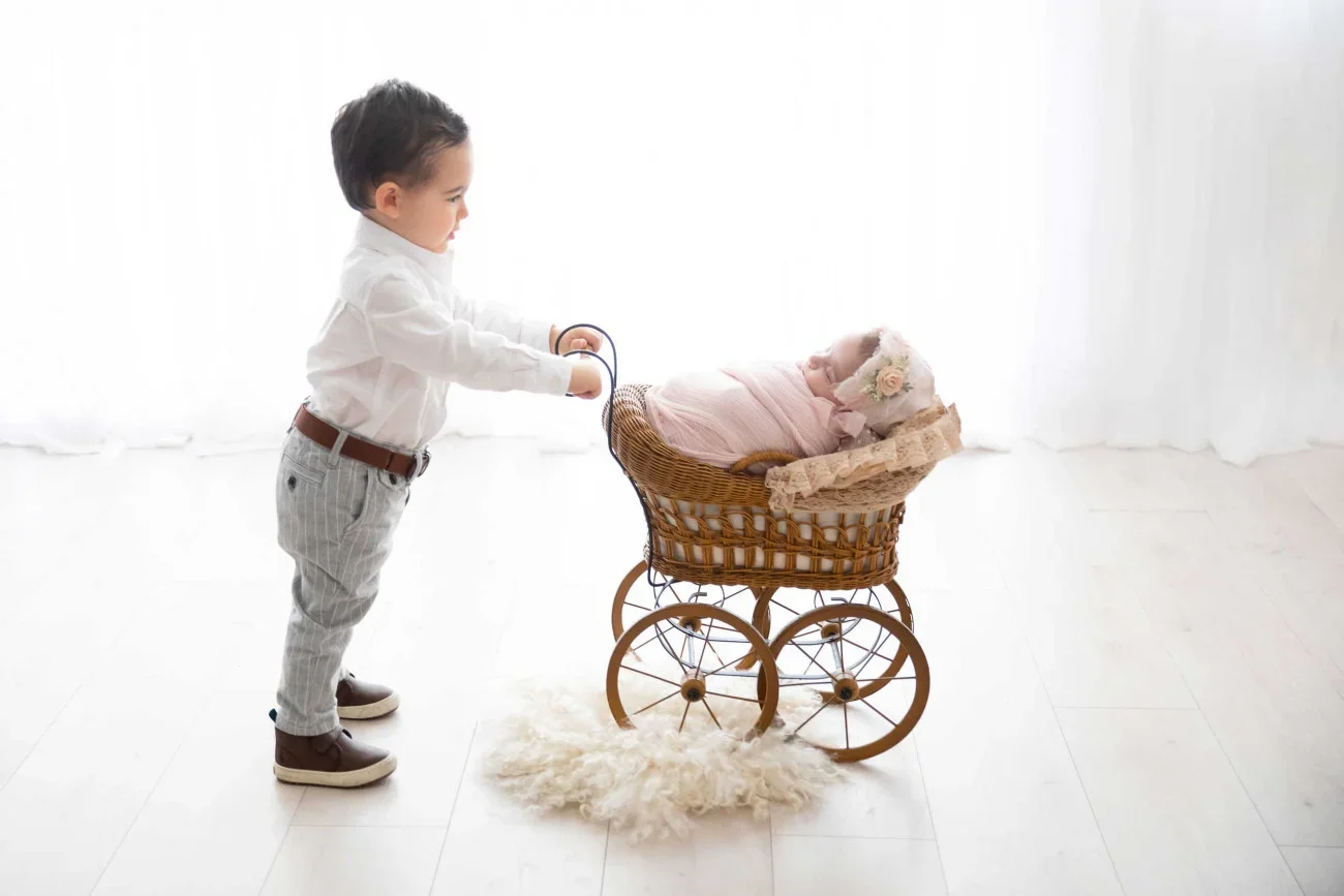 A young boy dressed in a white shirt and gray striped pants pushing a vintage doll stroller with a sleeping baby doll inside, covered with pink blankets, in a bright, white room.