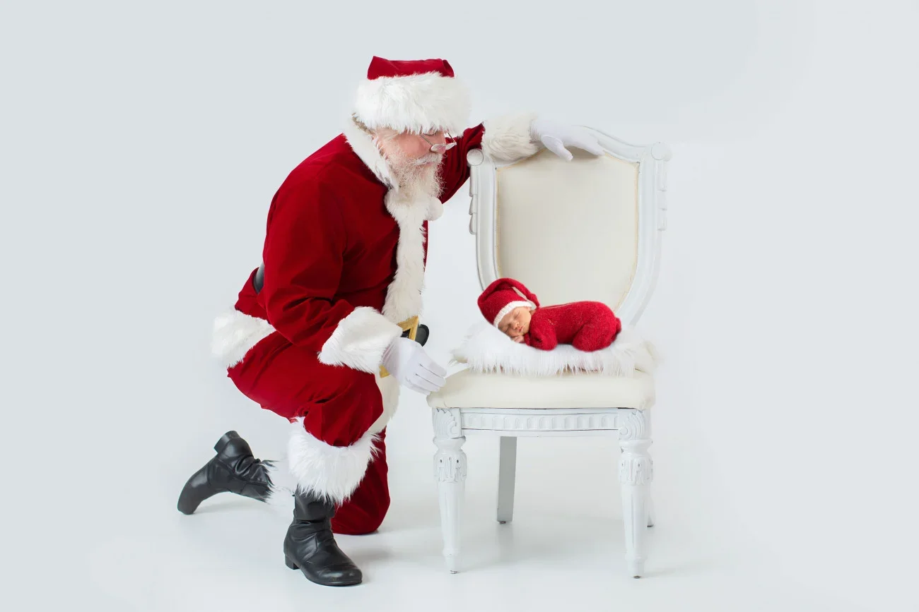 Santa Claus dressed in a red suit with white fur trim kneeling next to a white ornate chair. A sleeping baby in a red outfit with a matching hat is lying on the chair, with a white fluffy throw underneath.