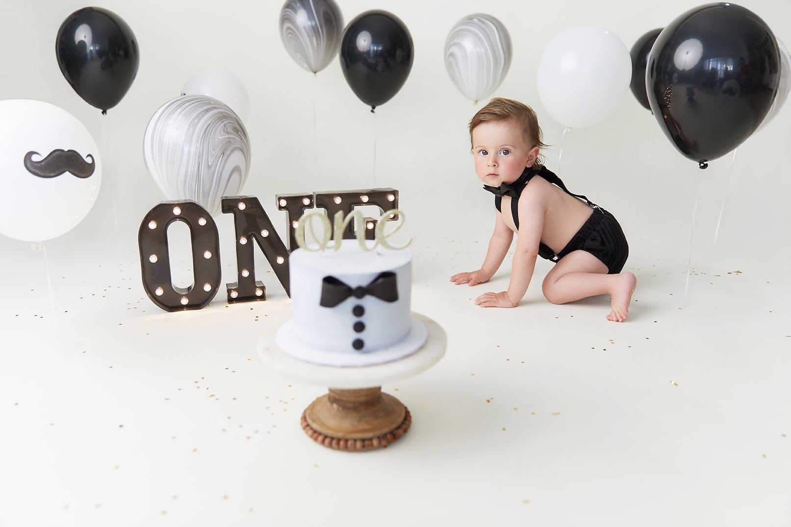Baby dressed in black bowtie outfit, crawling on white floor, surrounded by black and white balloons, a 'ONE' sign, and a small cake with a tuxedo design for a first birthday celebration.