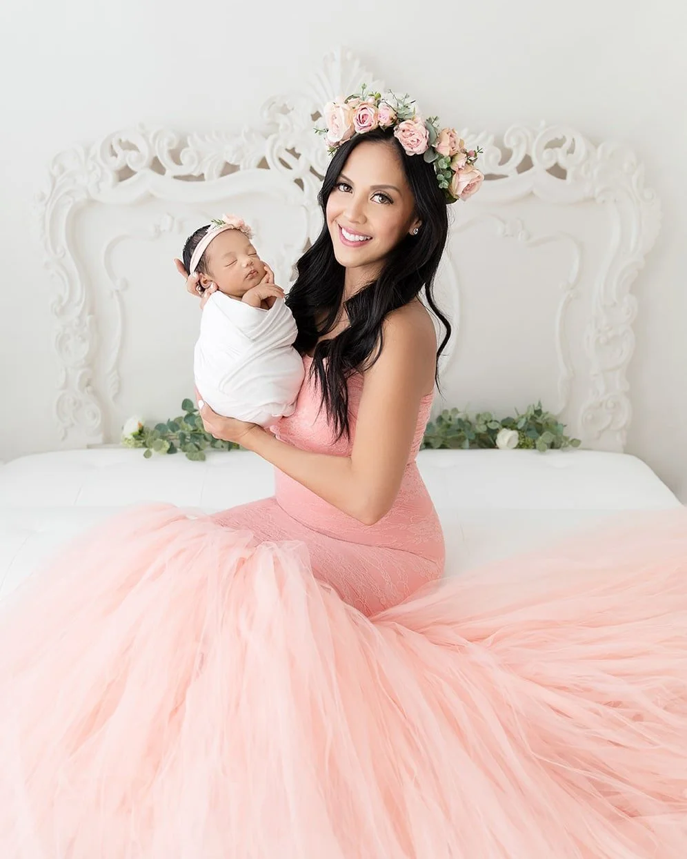 A woman with long black hair wearing a pink dress and a flower crown smiling while holding a sleeping baby wrapped in a white blanket, sitting on a white bed with ornate headboard and green and white floral decorations.