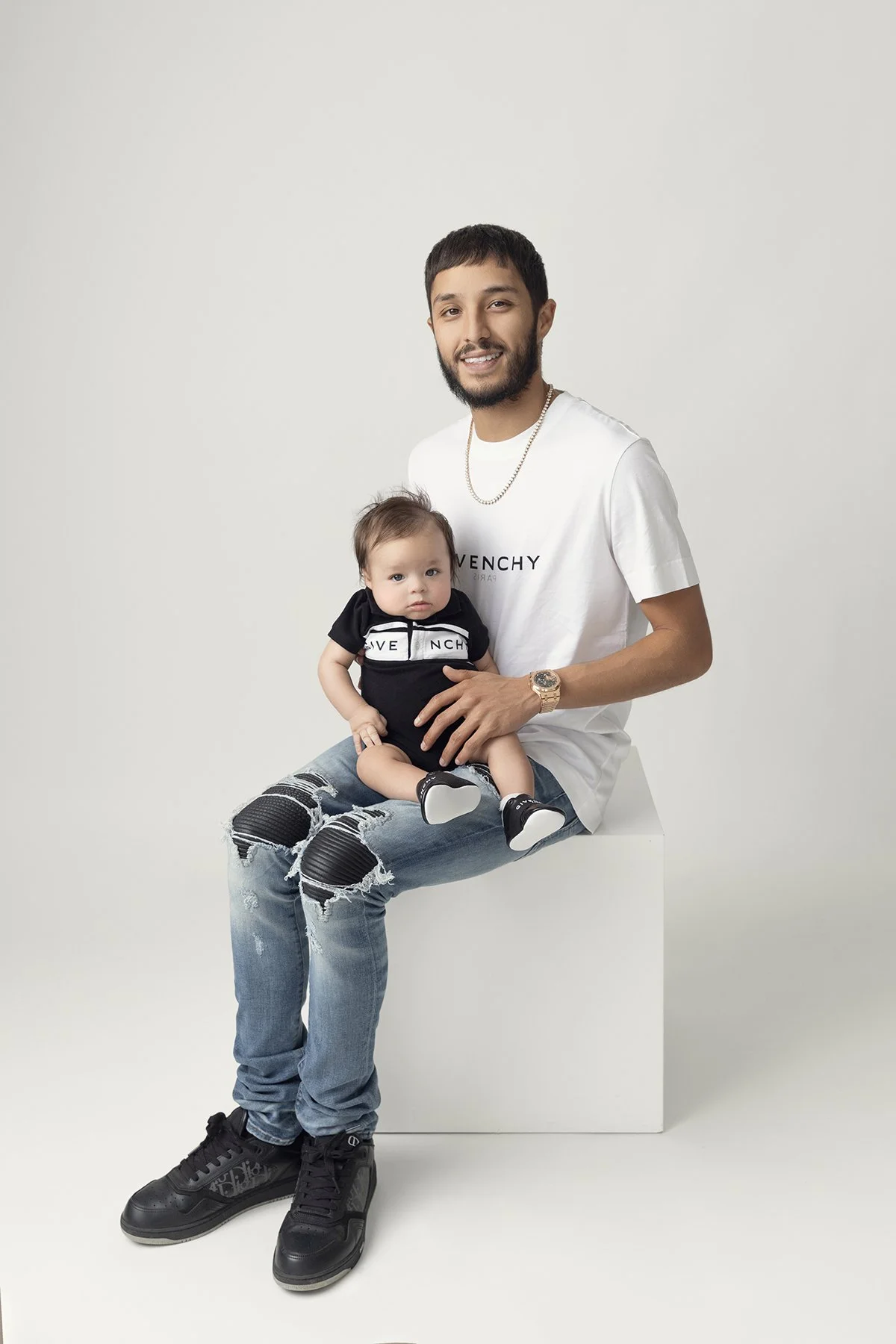 A smiling man with a beard, wearing a white Givenchy t-shirt, ripped jeans, black sneakers, a watch, and a chain, sitting on a white pedestal. He is holding a baby dressed in a black Givenchy outfit, with shoes, sitting on his lap against a plain whi