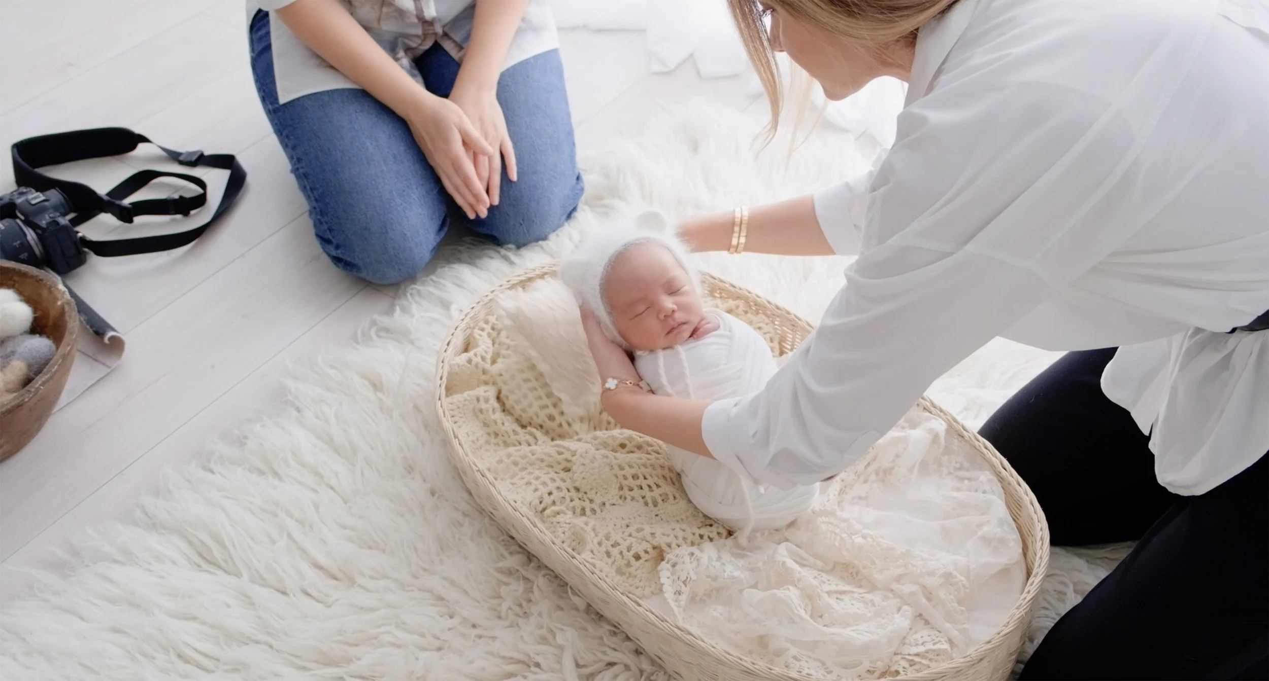 A newborn baby dressed in white lying in a woven bassinet on a soft white rug, while a woman adjusts the baby's head and a person kneels beside them. A woman sits nearby on the floor, and a camera and a basket of small items are on the floor.