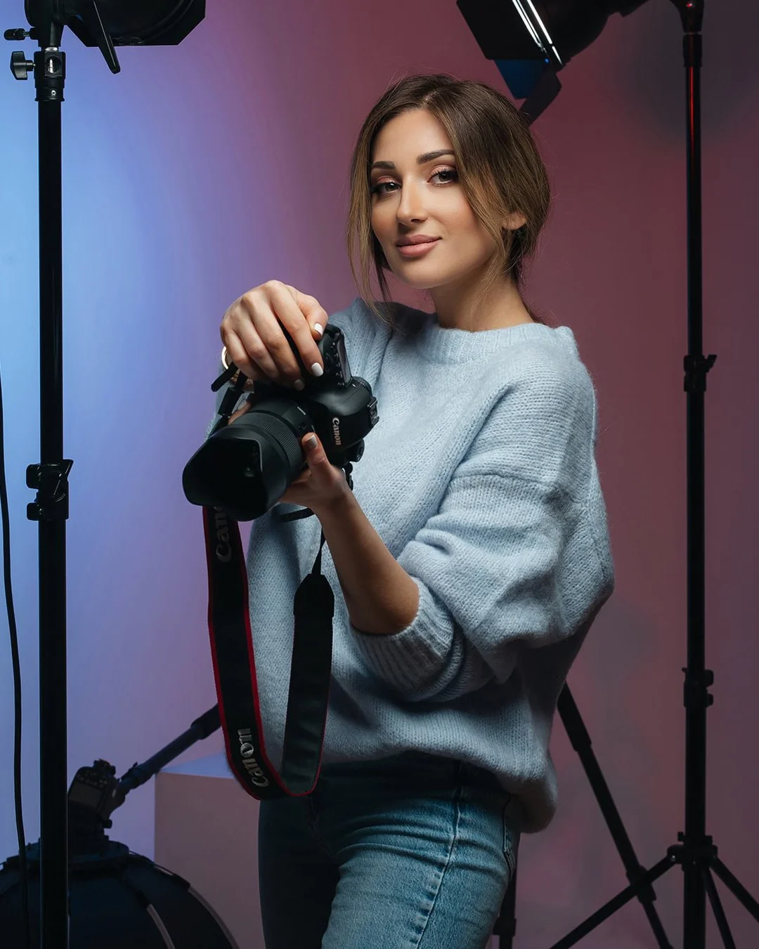 A woman with shoulder-length brown hair holding a Canon DSLR camera, standing in a photography studio with professional lighting equipment around her, wearing a light gray sweater.