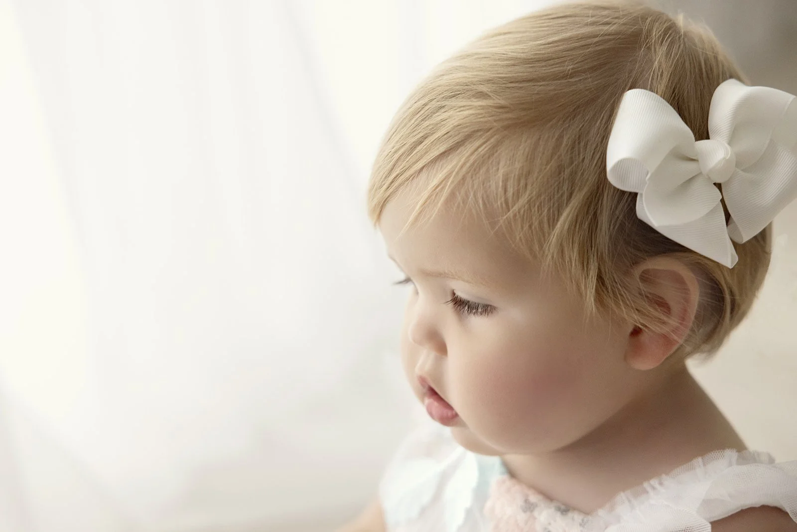 Profile of a young girl with short blonde hair wearing a large white bow, looking downward with a calm expression, softly lit background.