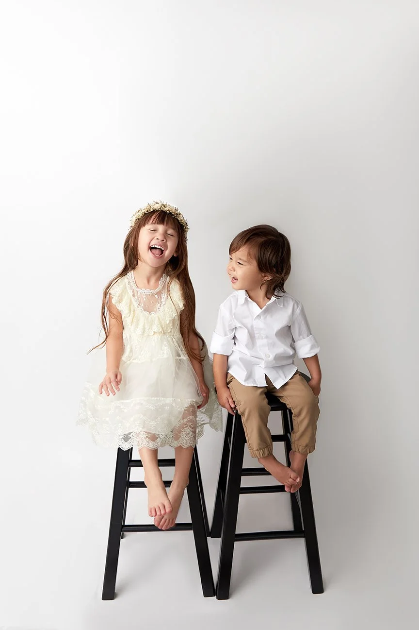 Two young children, a girl and a boy, sitting on black stools against a plain white background, laughing and smiling.