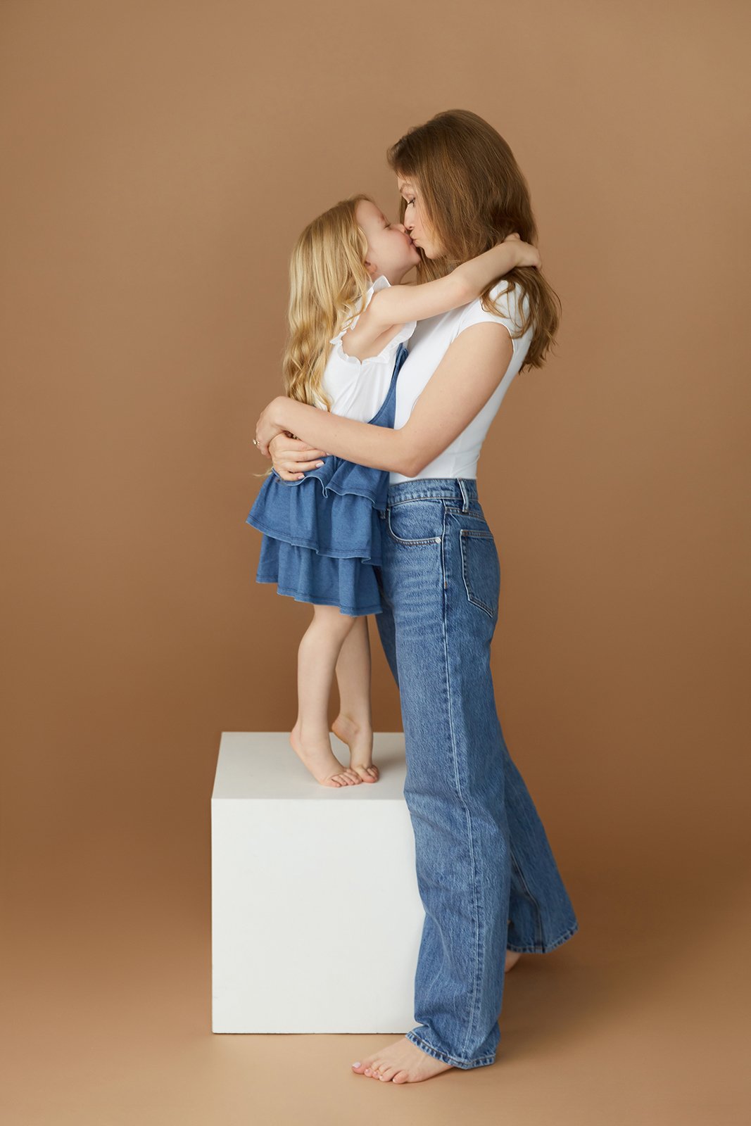 A woman and a young girl share a kiss, standing against a plain brown background. The girl is perched barefoot on a white cube, wearing a white top and blue skirt, while the woman wears a white t-shirt and blue jeans.