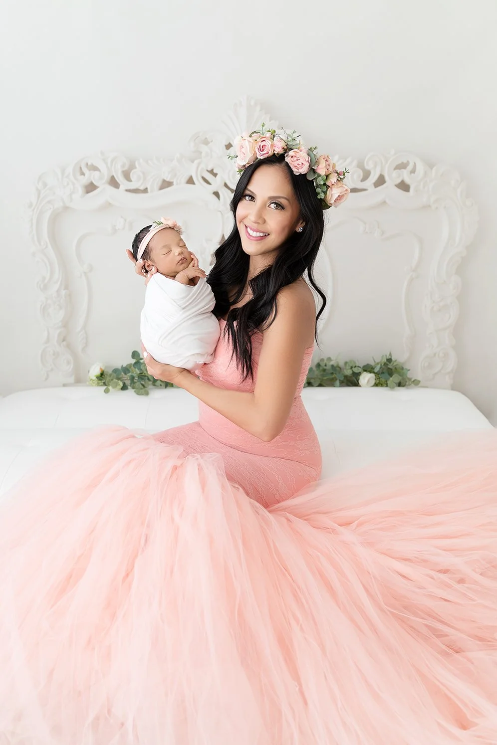 A woman with long dark hair wearing a pink gown and a floral crown holds a sleeping newborn wrapped in white while sitting on a bed with a white ornate headboard and green foliage decoration.