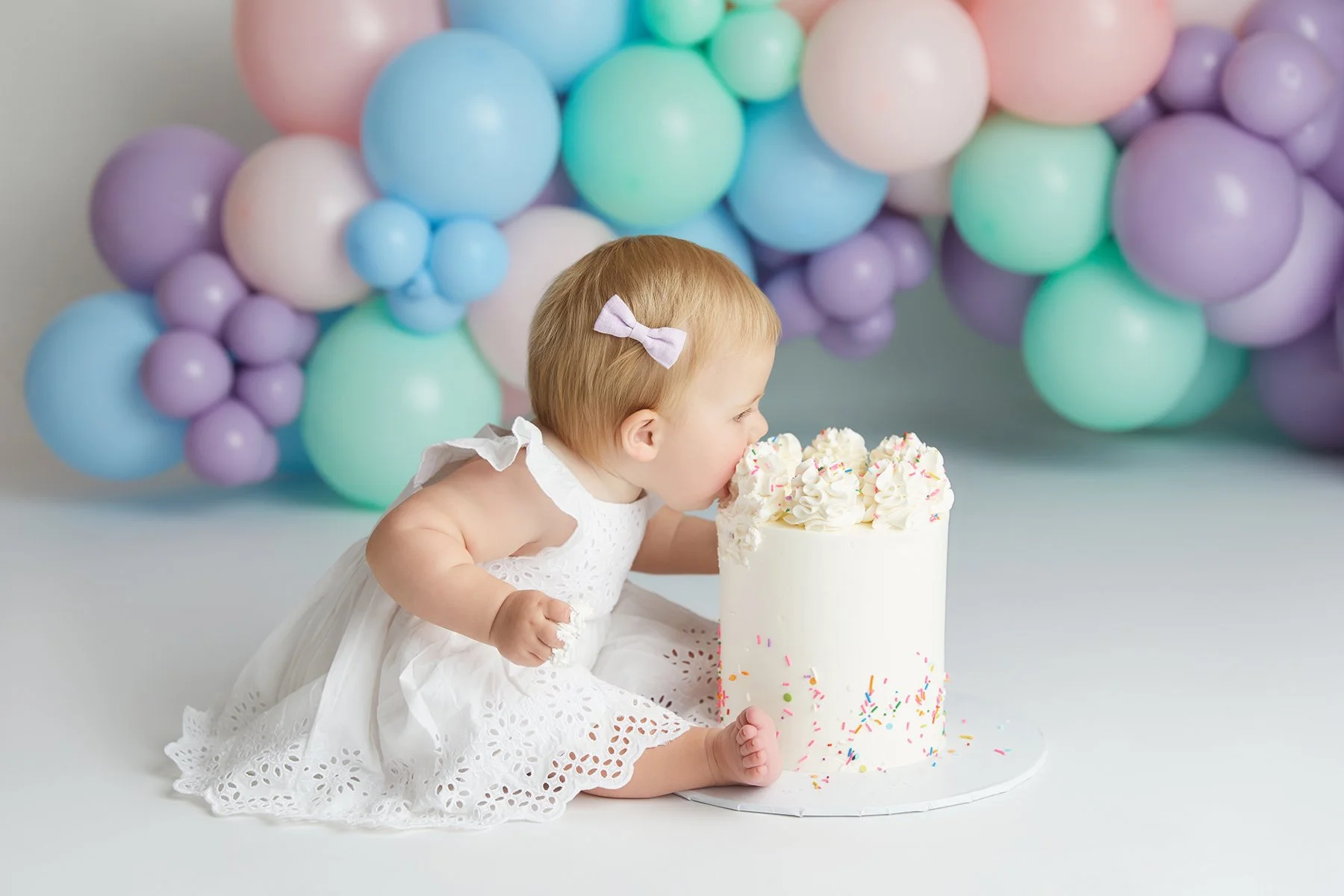 A young child with a pink bow in her hair wearing a white dress, leaning forward to blow out the candles on a tall white birthday cake decorated with sprinkles and frosting, with colorful balloons in pastel shades in the background.