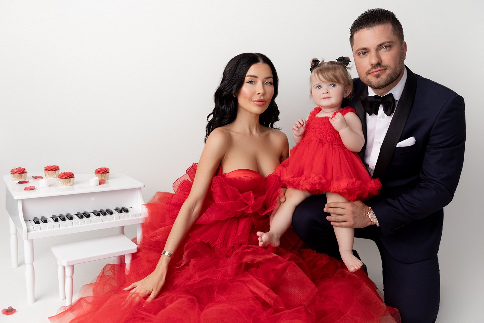 A family of three posing in front of a plain white background. The woman is wearing a large, red, strapless gown with a voluminous tulle skirt. She has black, wavy hair and is sitting on the floor. The man is dressed in a black tuxedo with a black bo