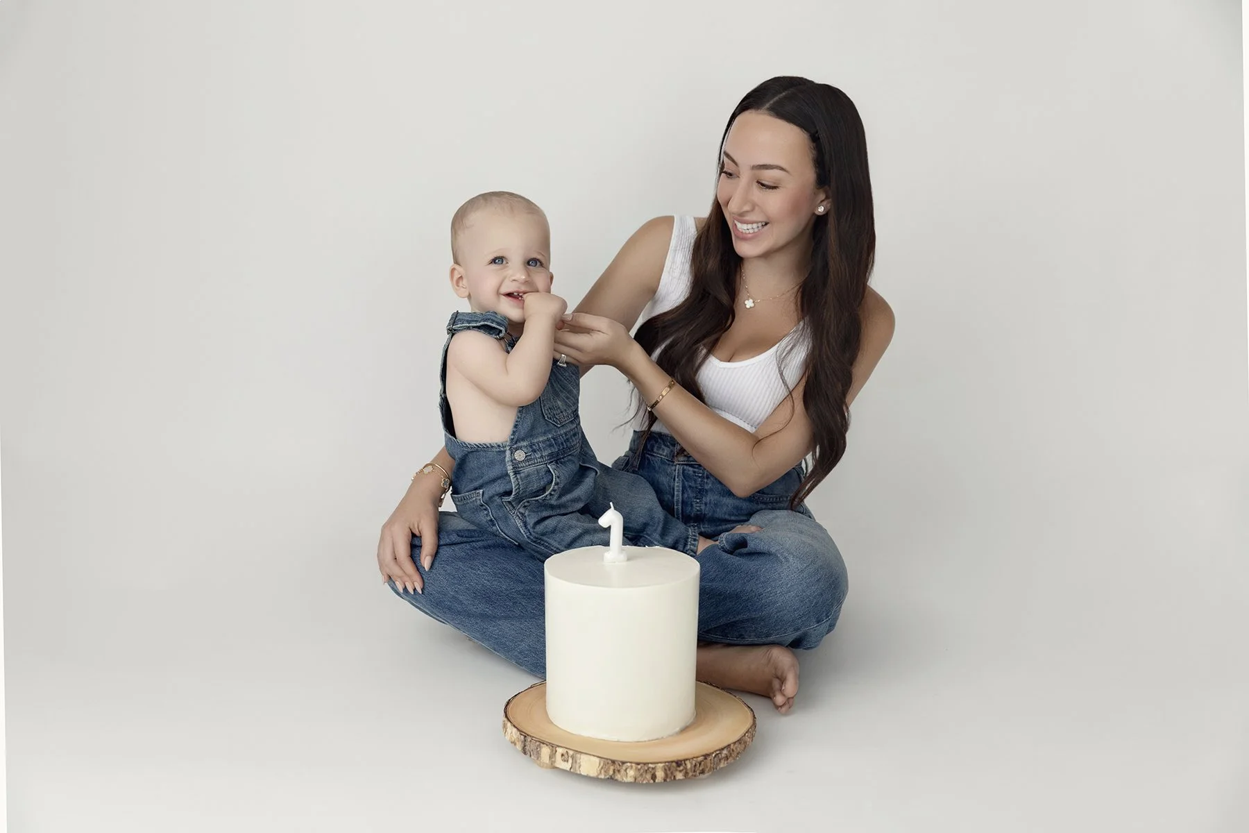 A woman and a young child sit on the floor with a white birthday cake with a single candle, smiling at each other.