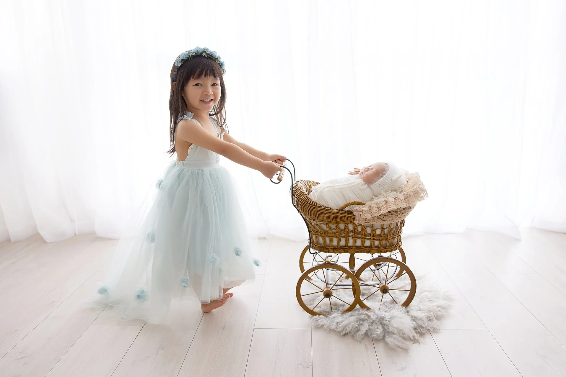 A young girl in a white dress with a floral headband stands barefoot, smiling, holding the handle of a vintage wicker doll carriage with a baby doll inside, against a white background.