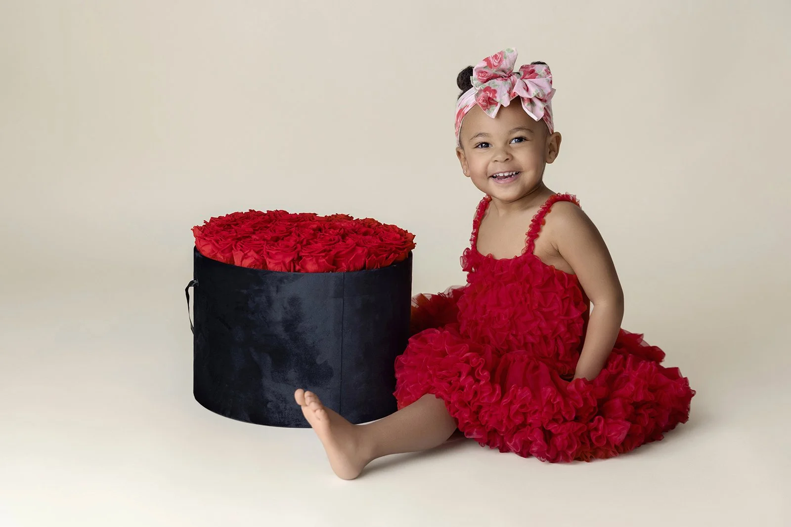 A smiling young girl wearing a red ruffled dress and a large pink floral headband sitting on the floor next to a round black box filled with red roses.