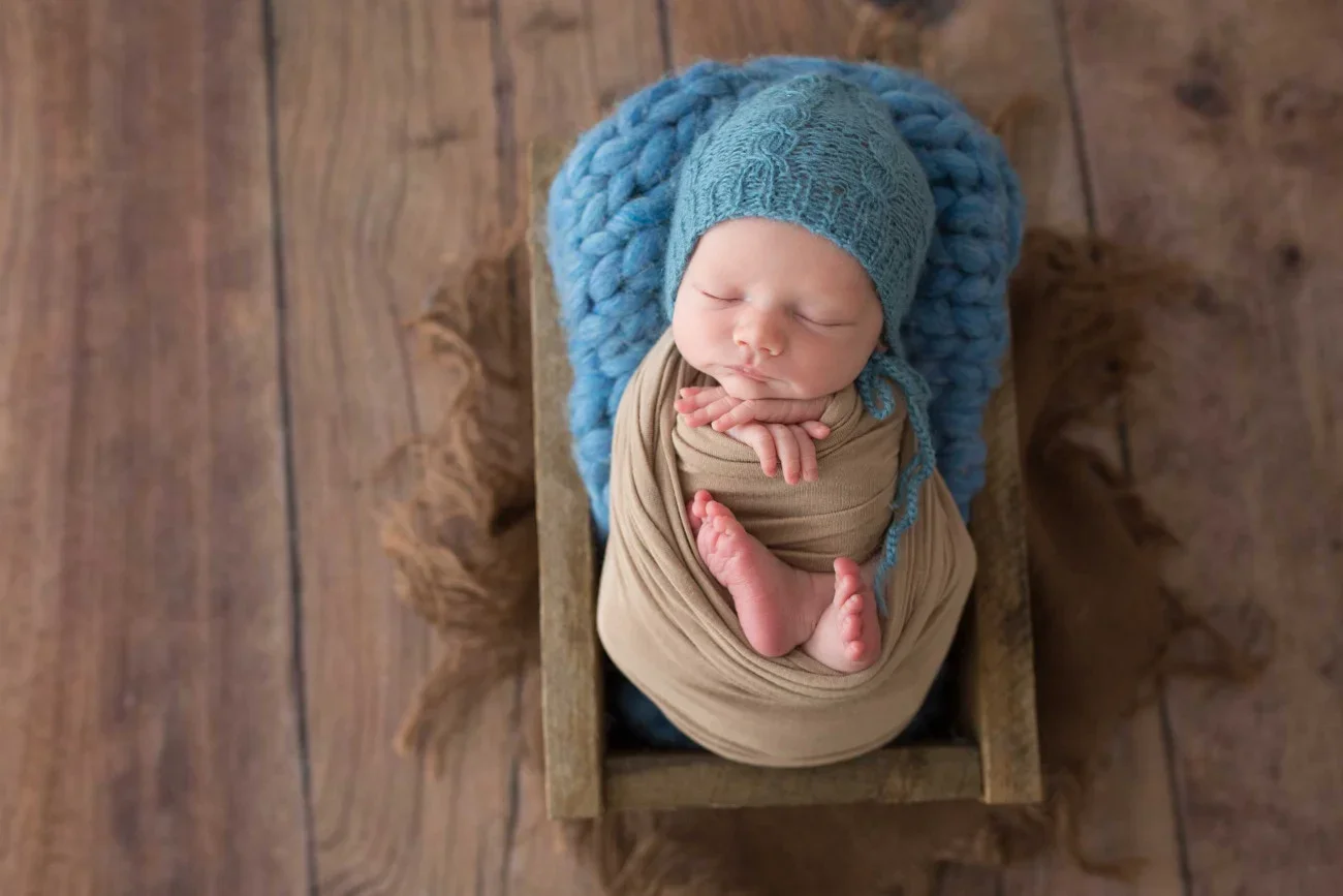 A newborn baby sleeping in a wooden box, wrapped in beige cloth, wearing a blue knit hat and bonnet, with a blue chunky knit blanket behind the head.