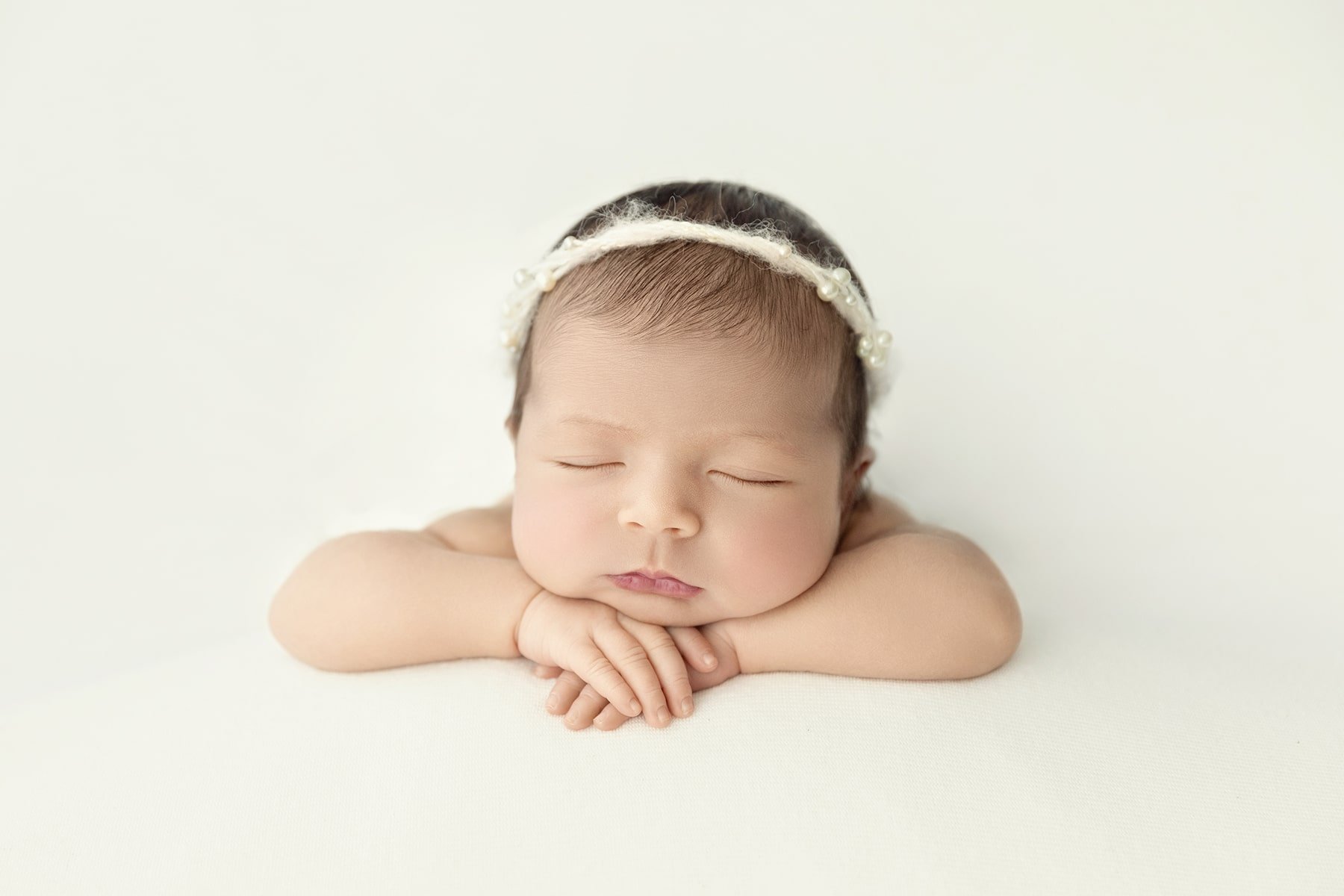 A sleeping baby with a pearl-embellished headband rests their chin on crossed arms against a white background.