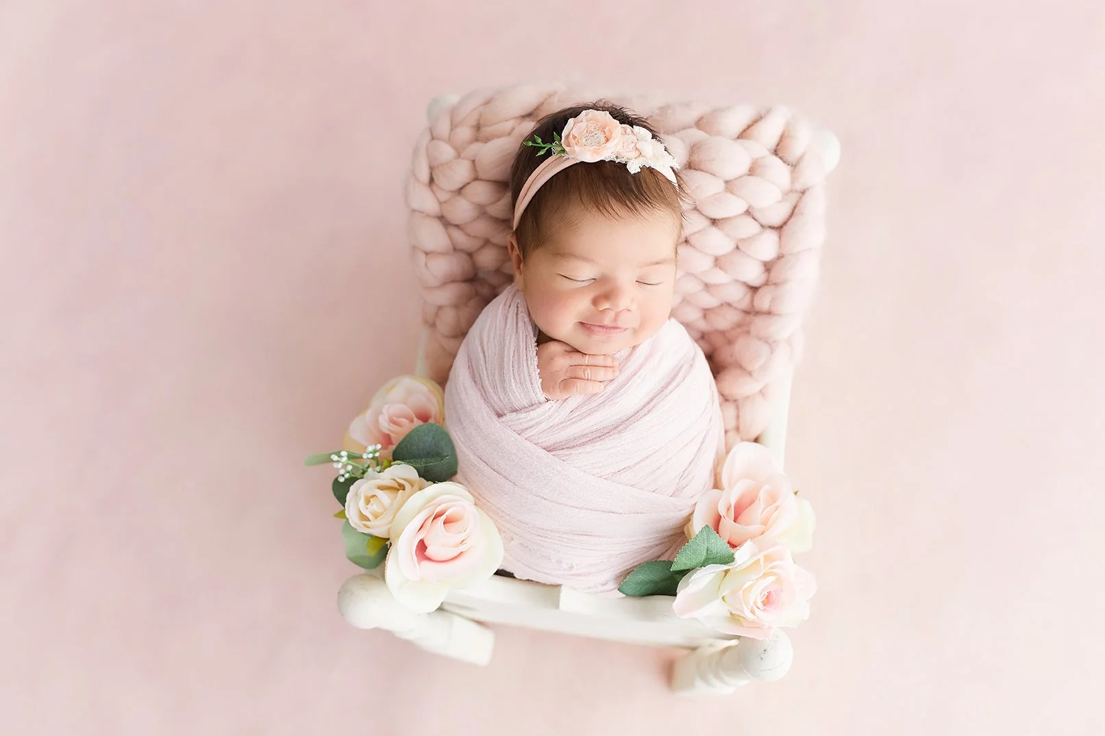 Young baby girl with a pink headband and flowers, sleeping in a white cradle decorated with pink and white roses, on a soft pink background.