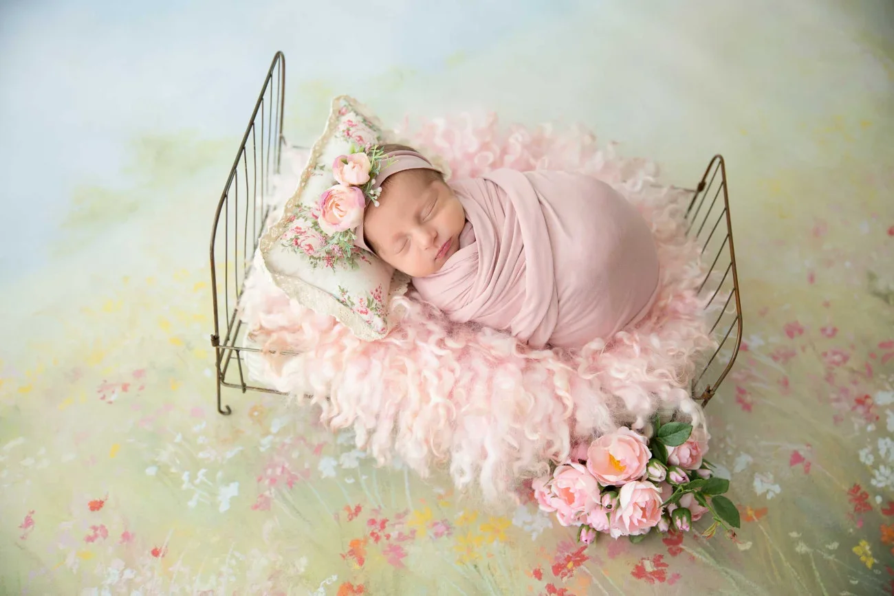 A sleeping newborn baby girl is wrapped in pink fabric, lying on a fluffy pink blanket inside a small wire bed. She wears a floral headband with pink roses, is resting on a pillow with floral embroidery, and is surrounded by pink roses. The backgroun