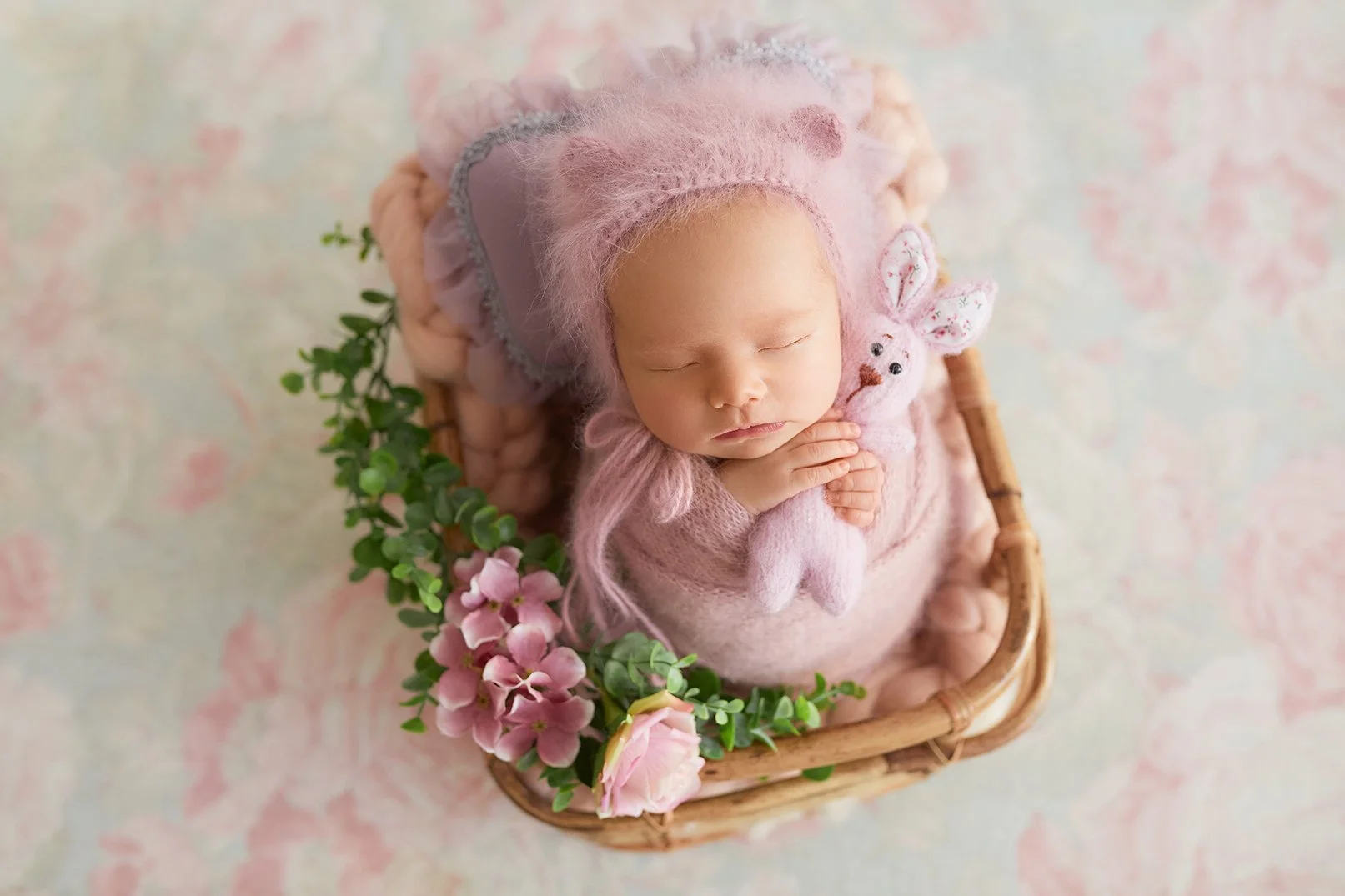 A sleeping baby in a pink fuzzy outfit holding a pink stuffed bunny, surrounded by pink flowers in a small wicker basket.