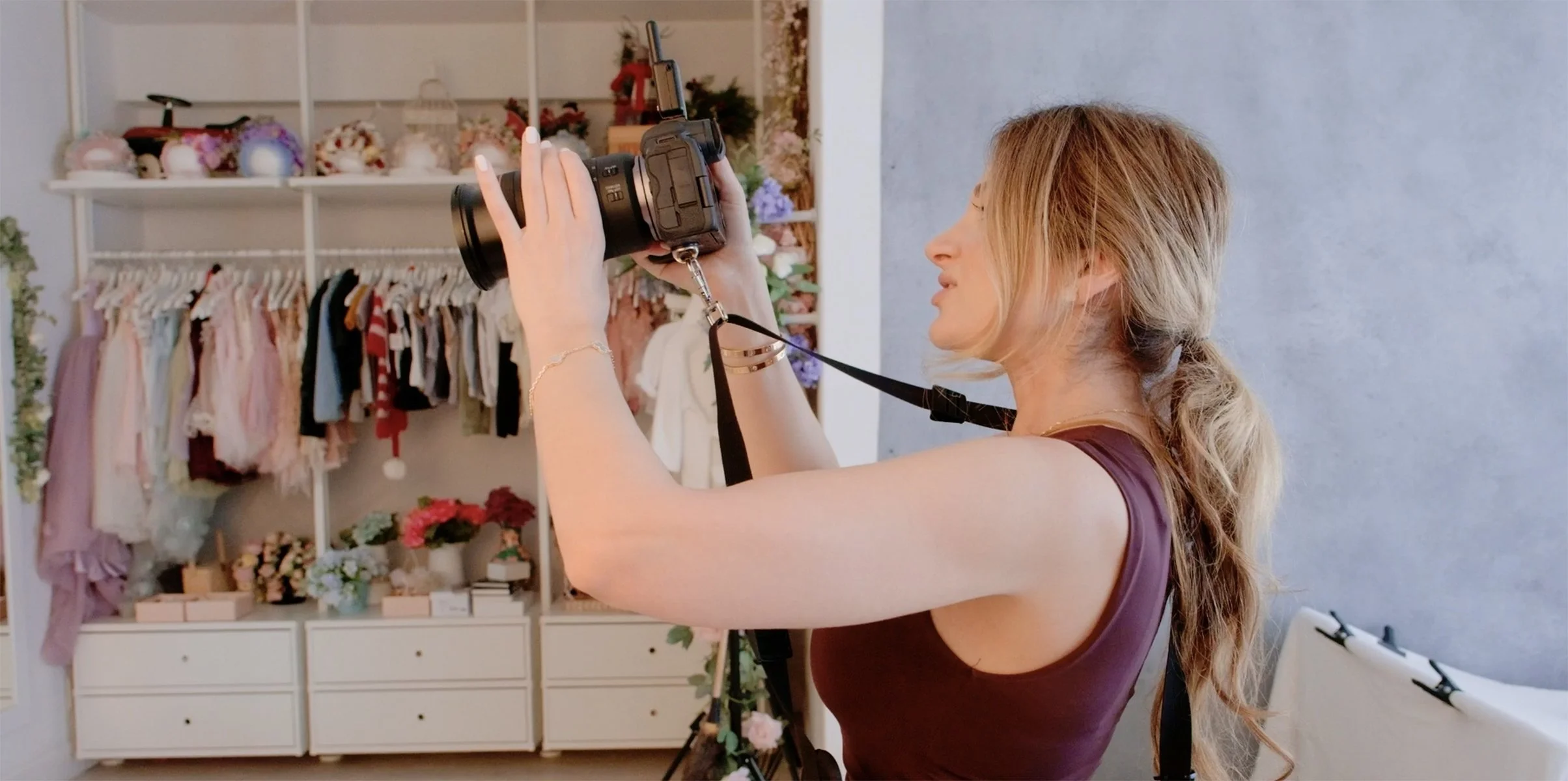 Woman taking a photograph indoors with a camera, standing in front of a shelving unit filled with clothes and decorations in a room with light-colored walls.