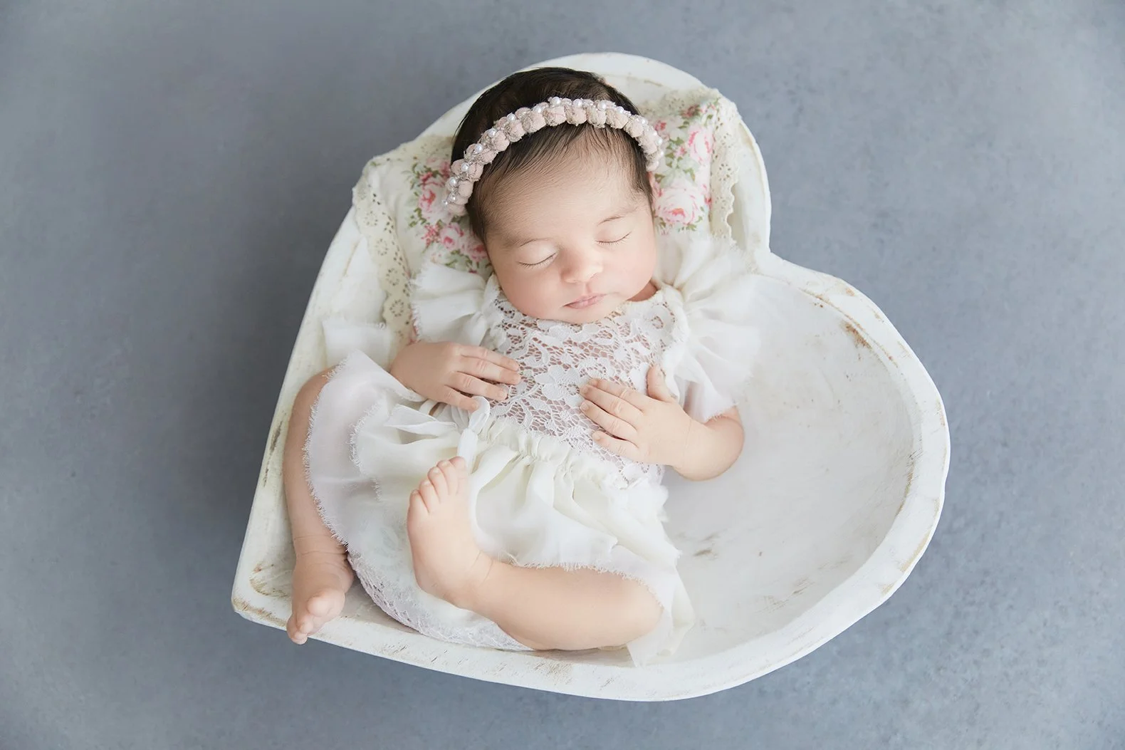 A sleeping baby girl in a white lace dress and a pearl headband, lying in a white bowl on a gray surface.