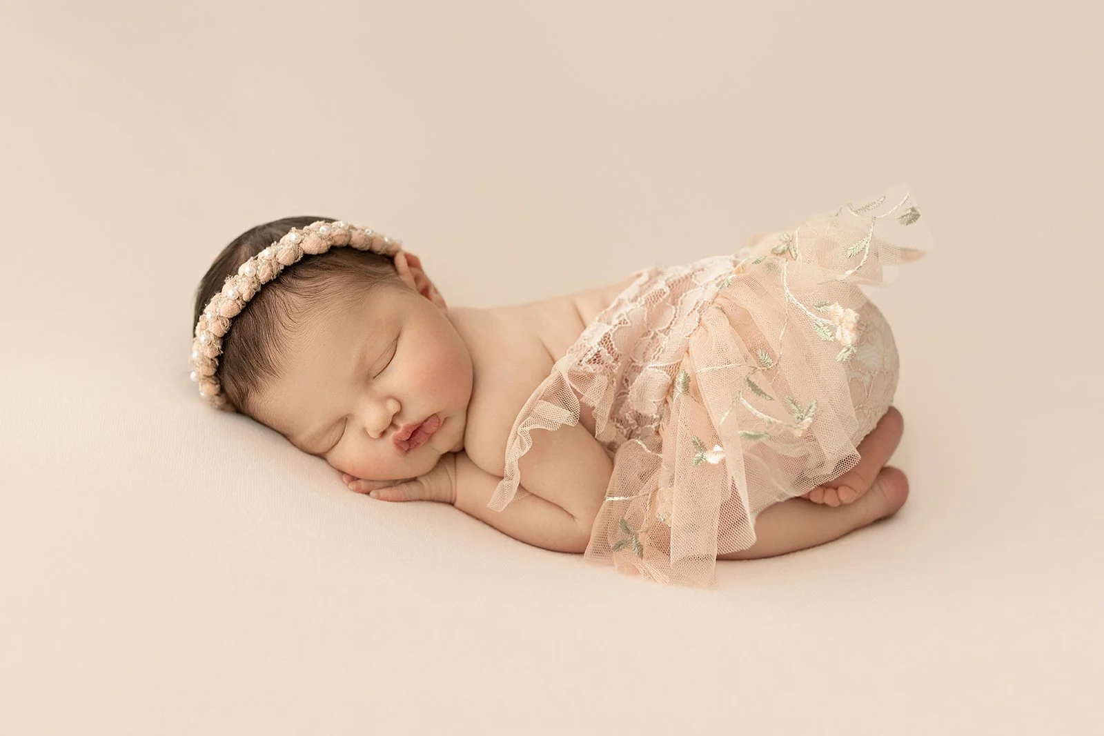 A sleeping baby girl lying on her side on a soft, light-colored surface, wearing a delicate pink lace dress and a floral headband.