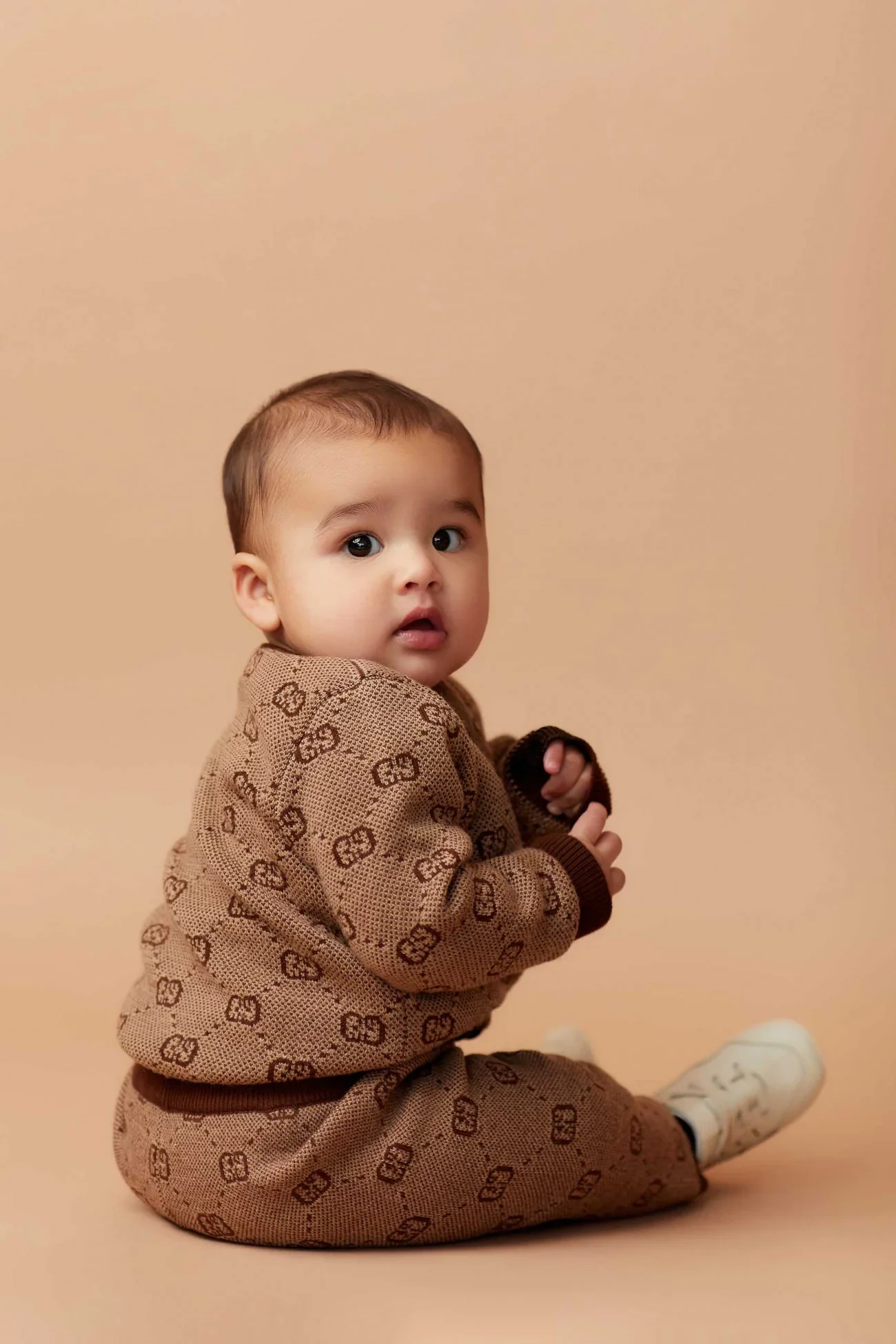 A young child sitting on the floor against a beige background, wearing a matching brown patterned outfit, and looking at the camera with a curious expression.