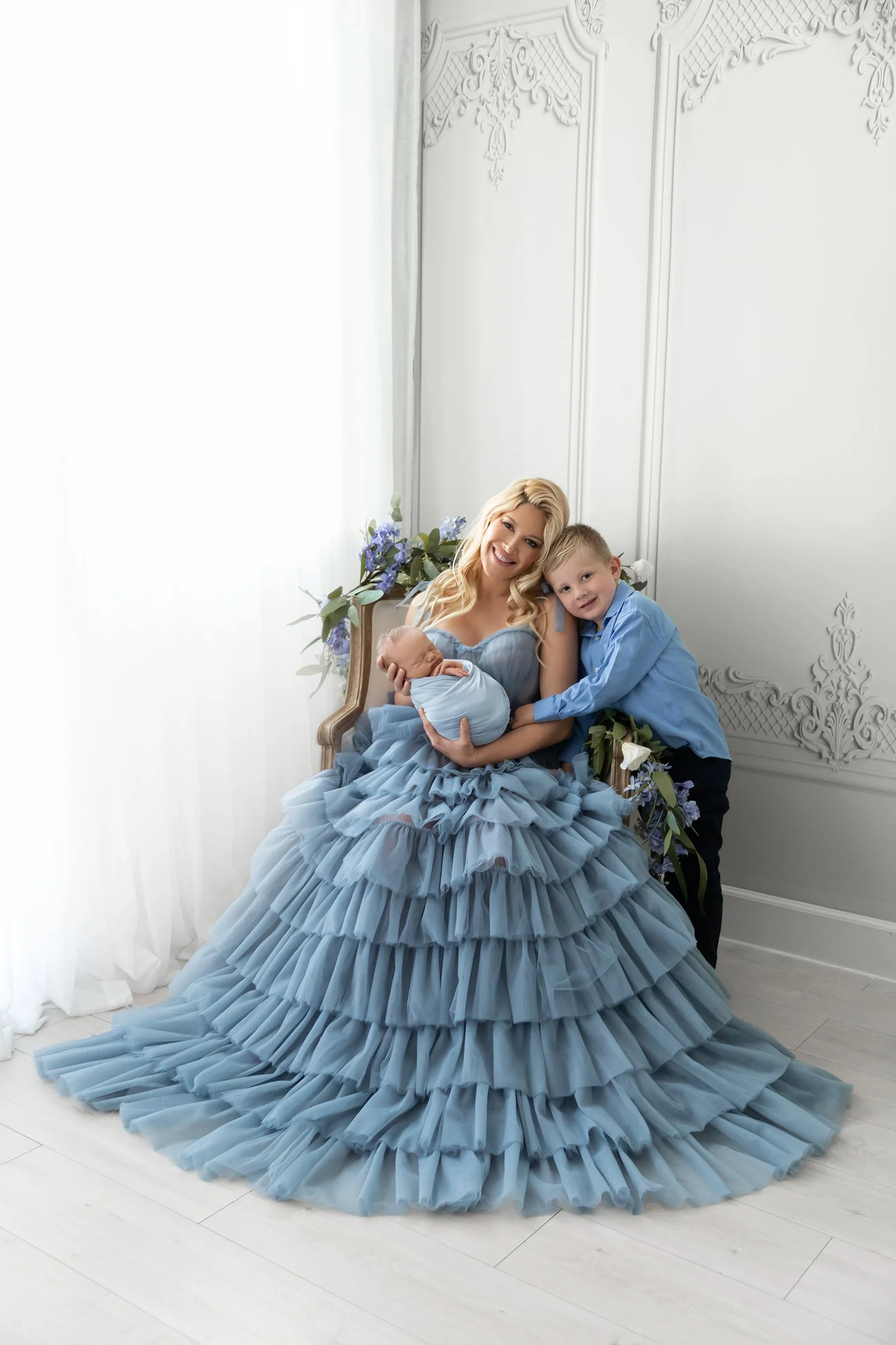 A woman in a ruffled blue dress sitting on a decorative chair, holding a newborn baby wrapped in a blue blanket, with a young boy leaning on her shoulder, surrounded by flowers against a white ornate wall.