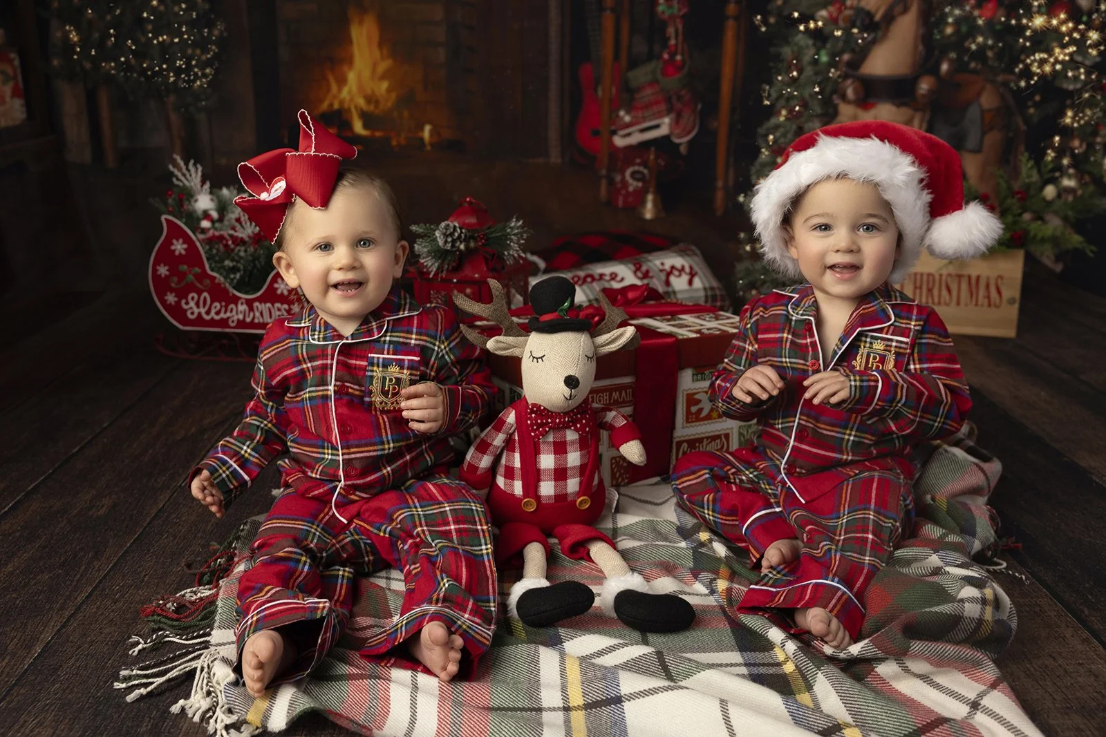 Two young children sitting on a plaid blanket in front of a decorated Christmas tree, surrounded by Christmas gifts and ornaments, dressed in red plaid pajamas with holiday accessories, one wearing a Santa hat and the other a large red bow, with a st