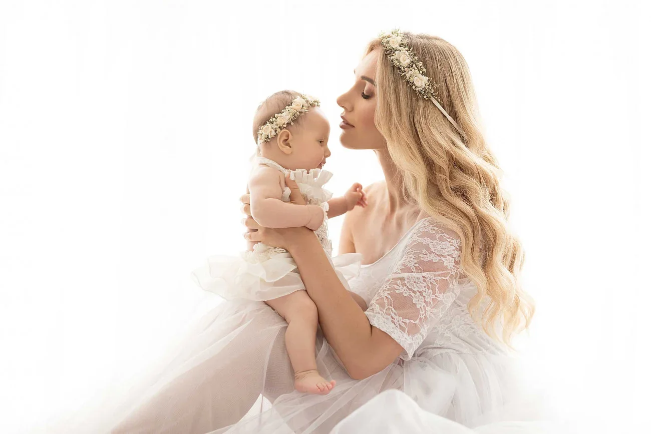 A woman with long blonde hair wearing a floral headband and white dress holding a baby girl dressed in white with a matching floral headband, against a bright white background.