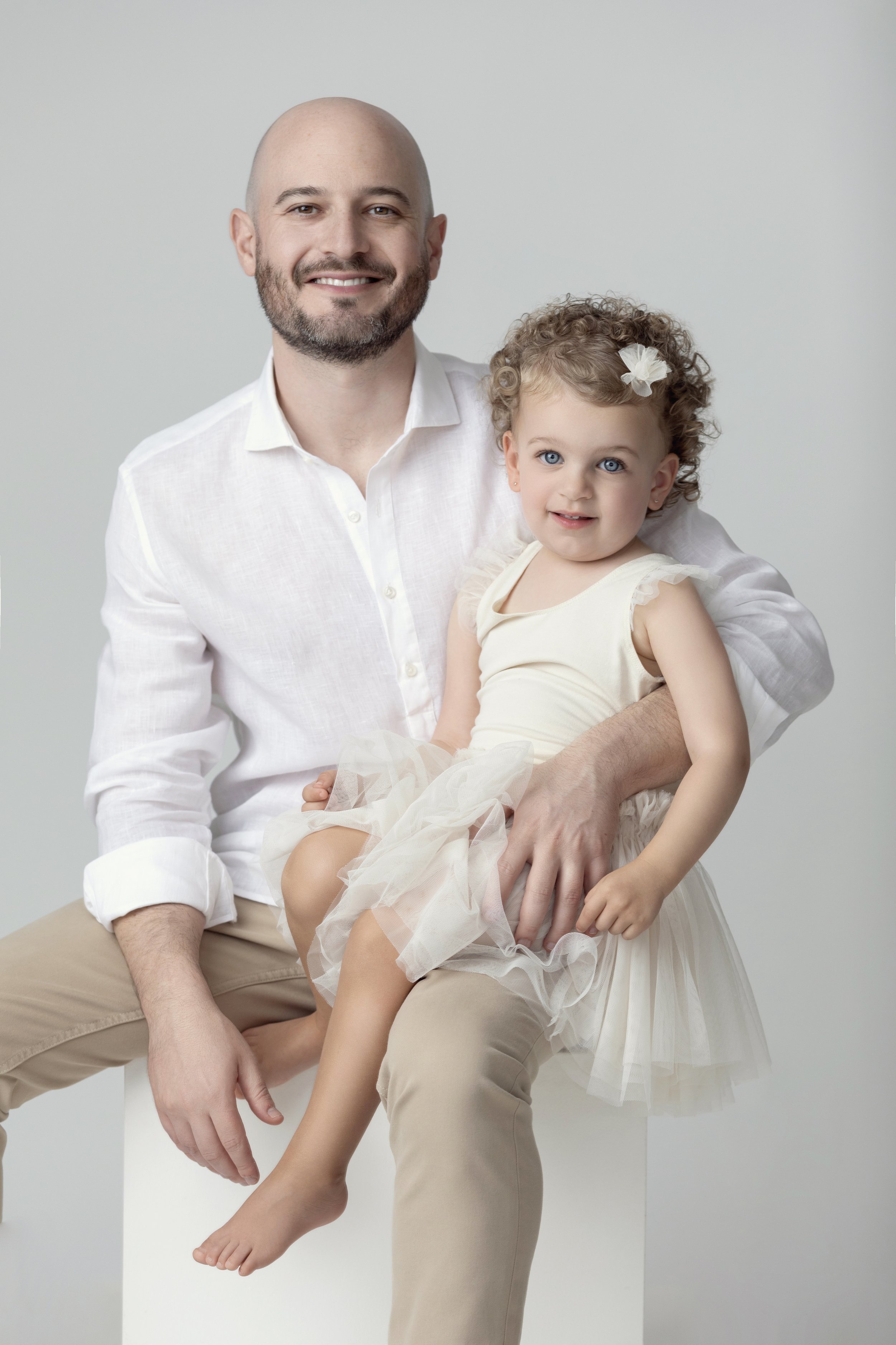 A man with a beard and a bald head, wearing a white shirt, holds a young girl with curly blonde hair, blue eyes, a white flower hair accessory, and a cream-colored dress with tulle. They are smiling against a plain light gray background.