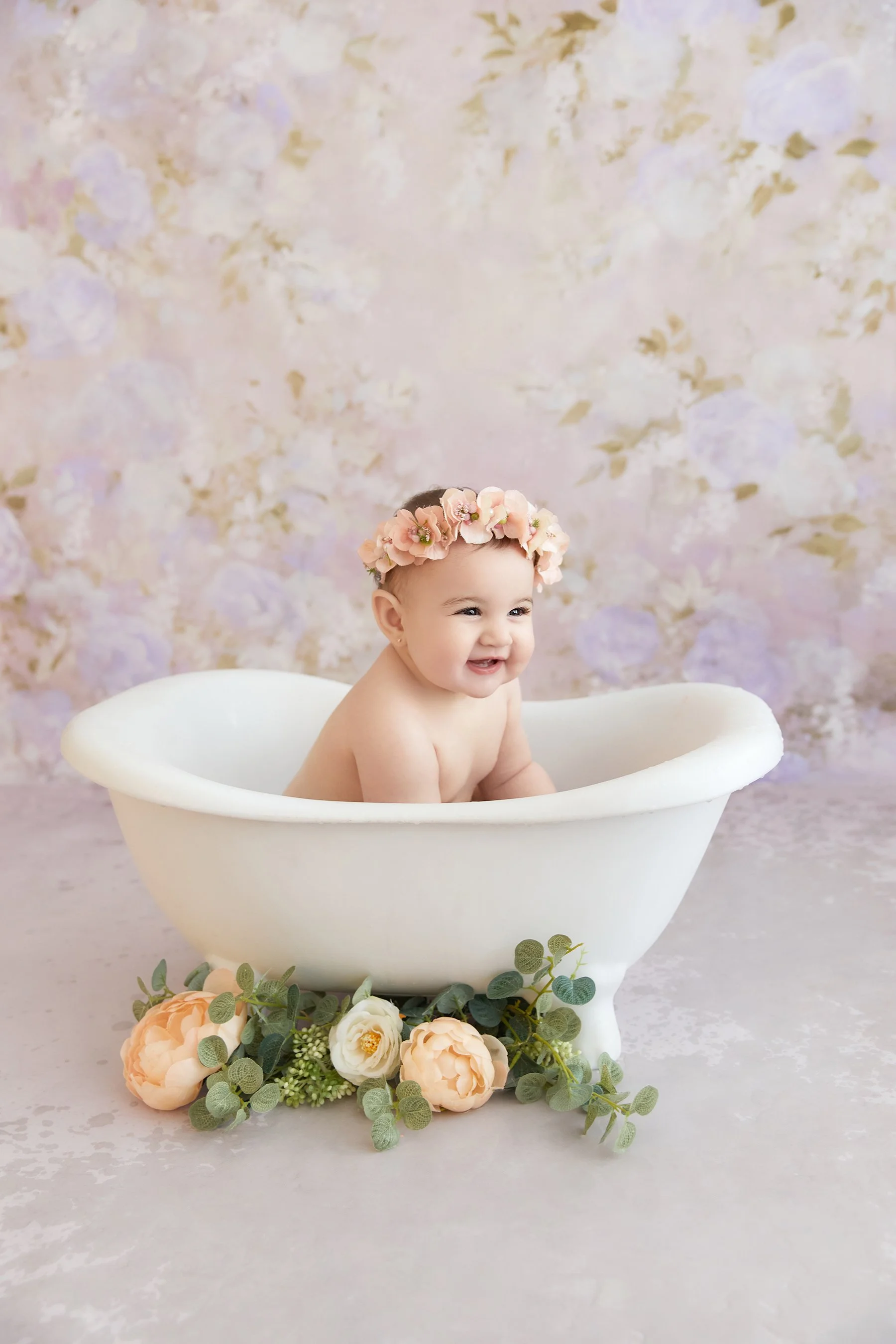 A smiling baby in a vintage-style white bathtub, wearing a pink floral crown, with pastel-colored flowers and greenery around the base of the tub against a soft floral backdrop.