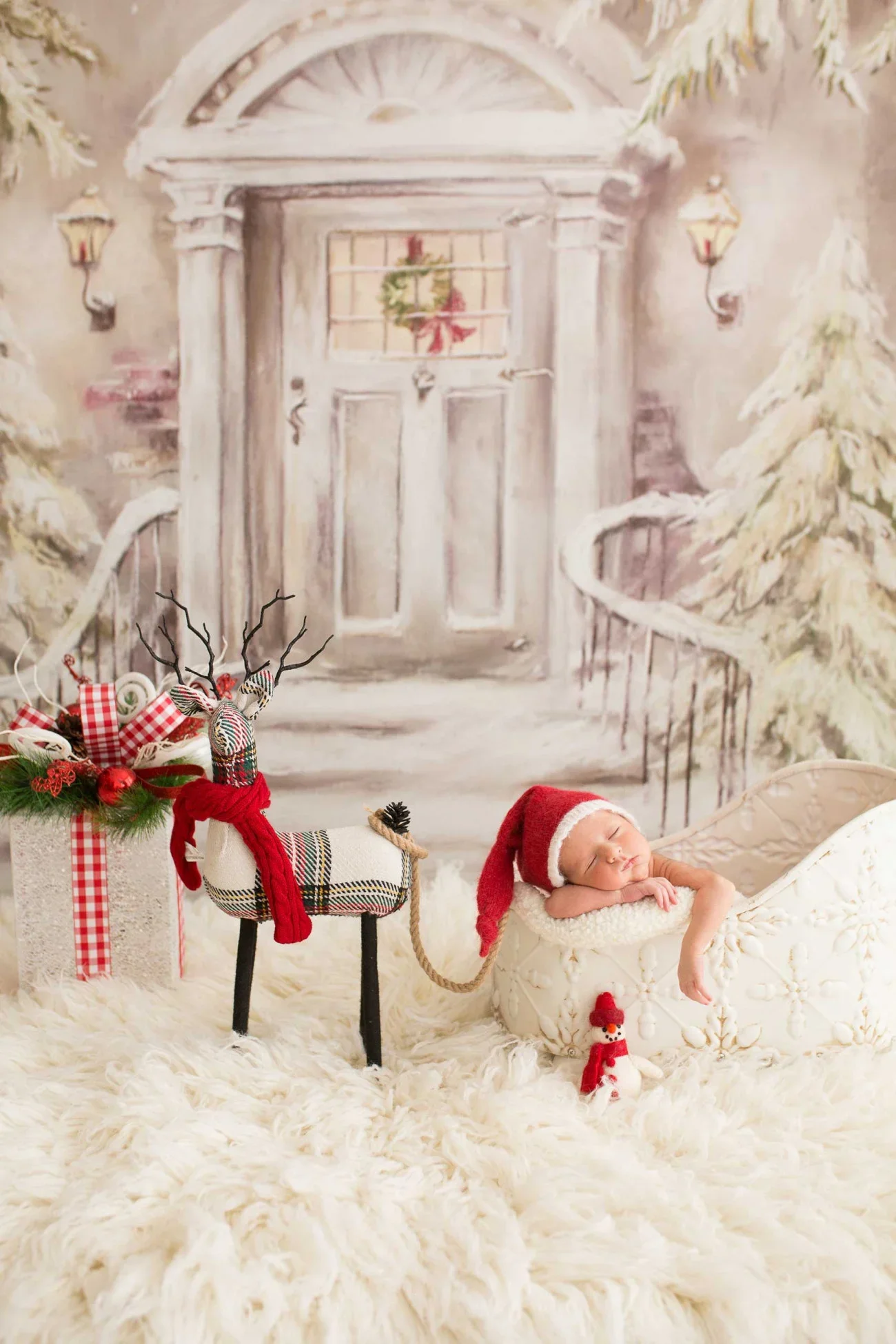 A sleeping baby wearing a Santa hat lying in a white vintage-style cradle with a small snowman figurine beside it. The scene is decorated with Christmas-themed items, including a reindeer figure wearing a red scarf, a festive arrangement with red orn